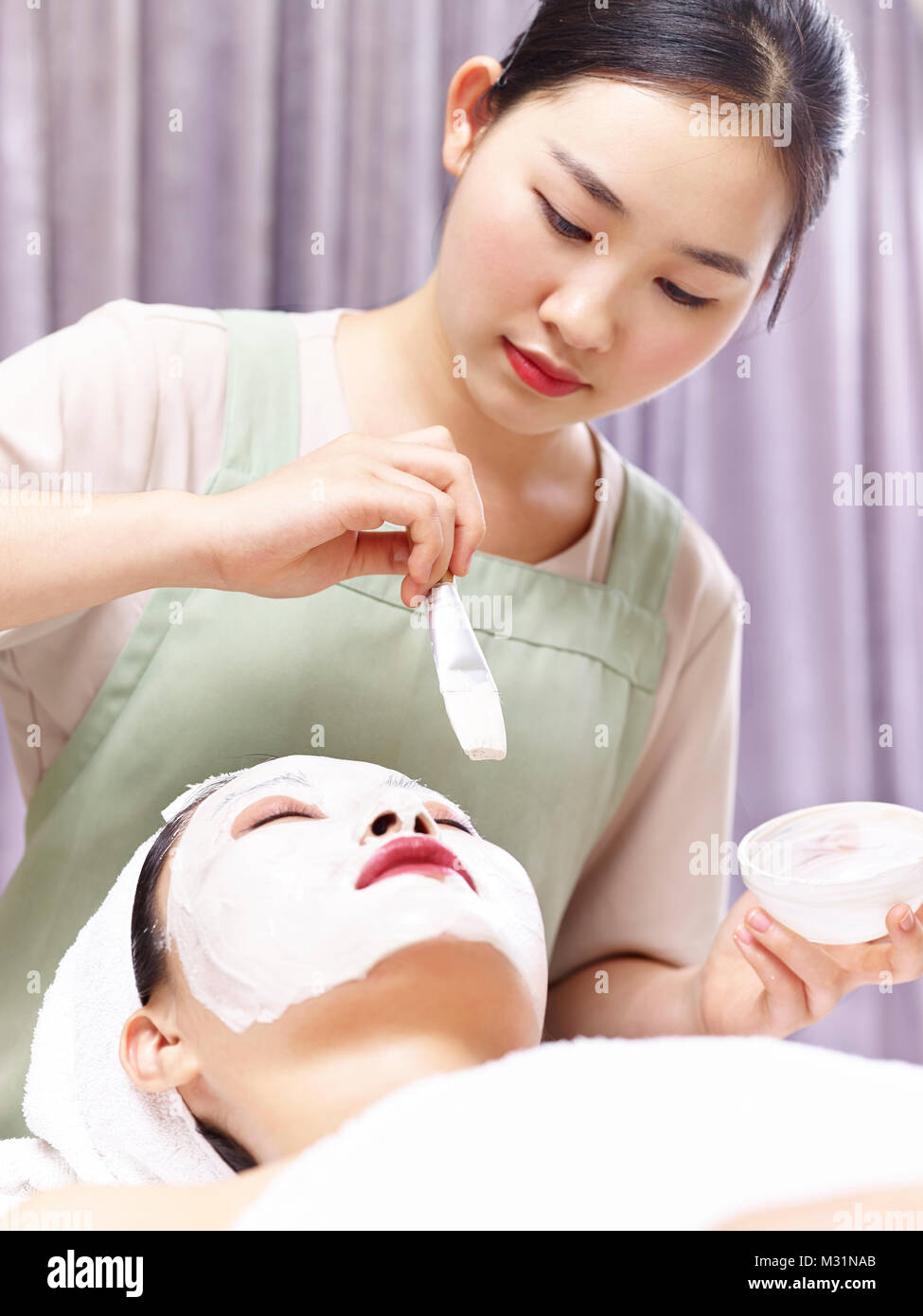 asian beautician applying white facial mask with a brush on face of a young woman. Stock Photo