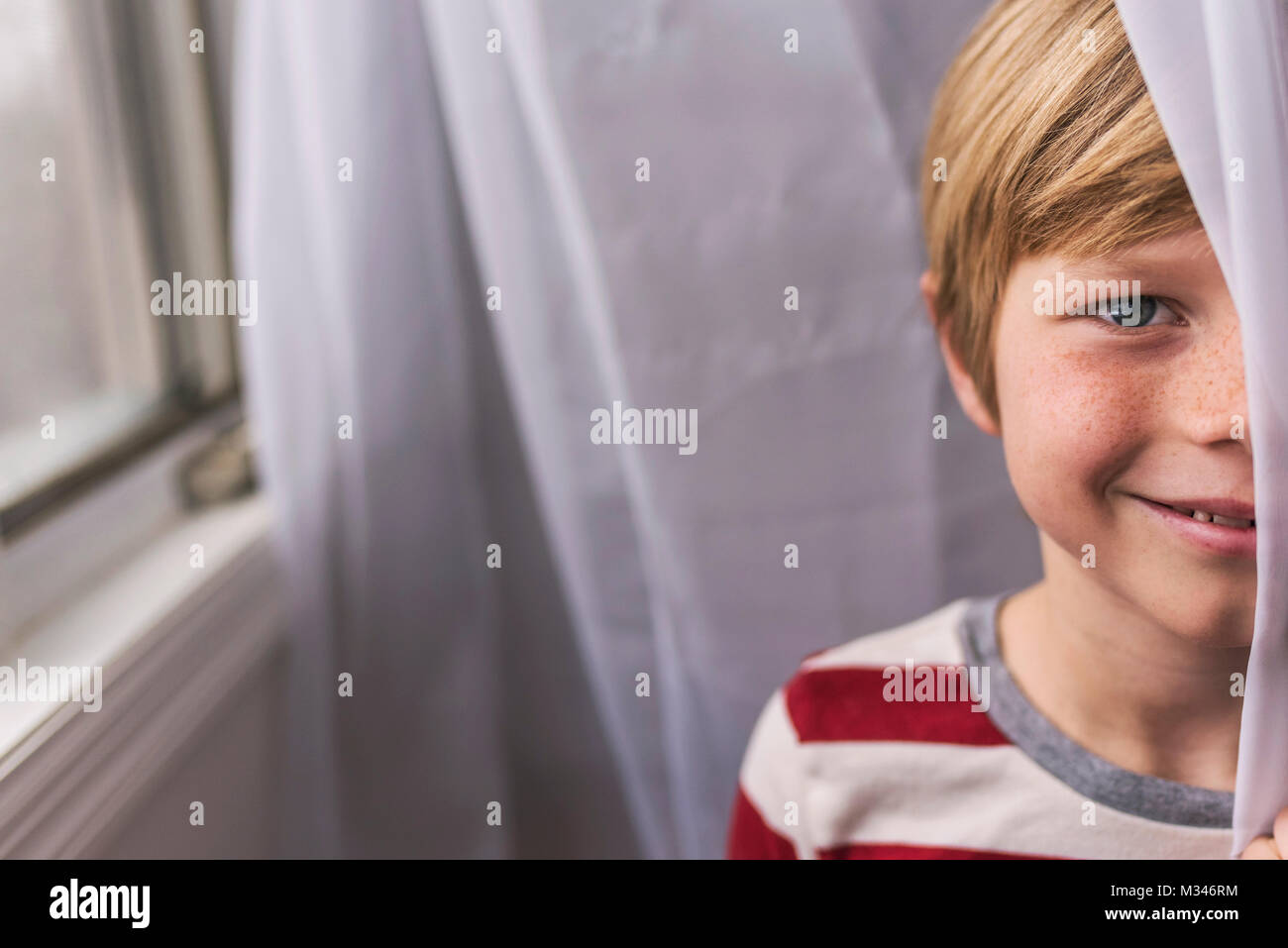 Portrait of a smiling boy with freckles standing by a window hiding behind a curtain Stock Photo