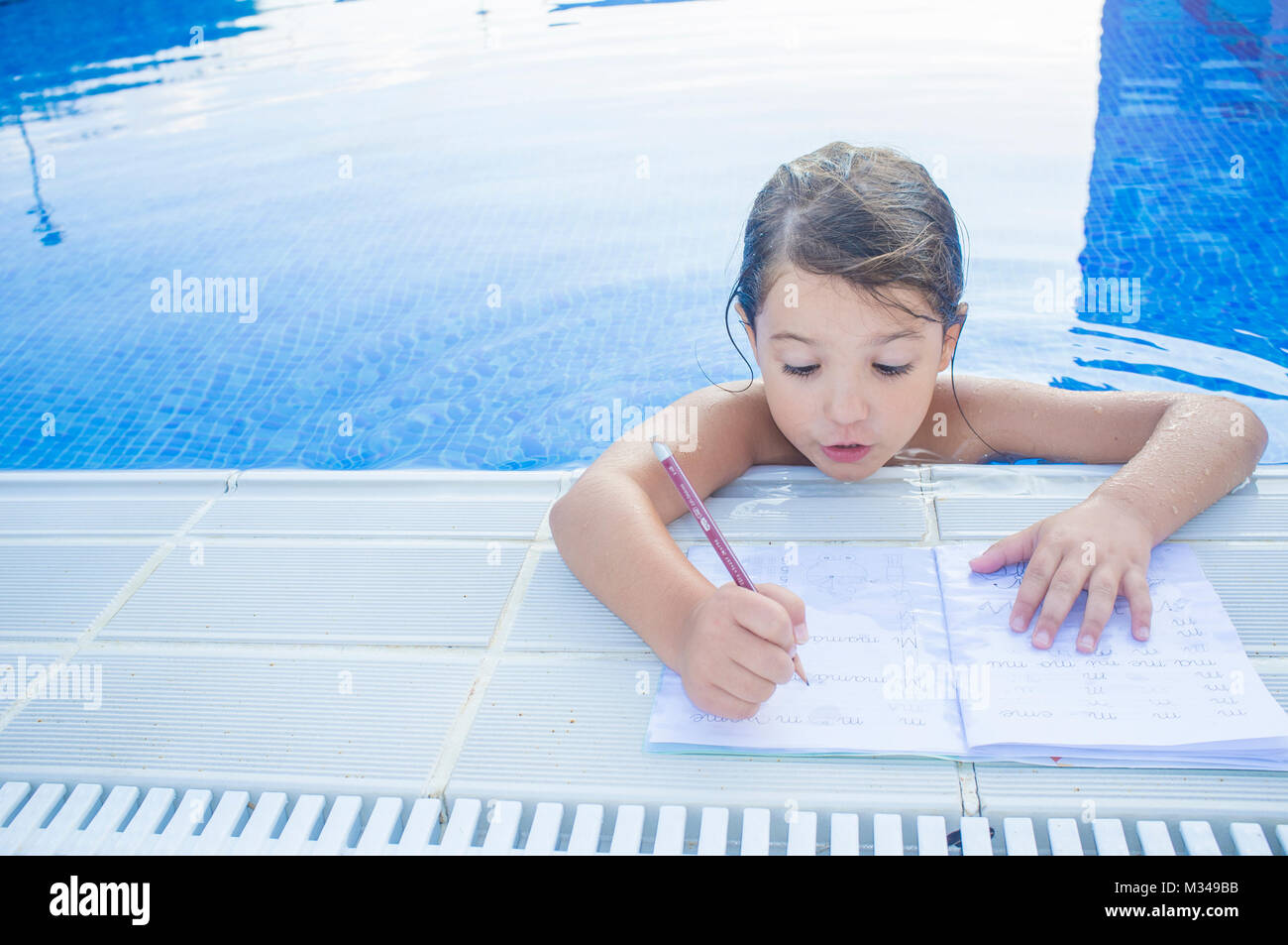 Child girl doing holidays homework over swimming poolside. Summer homework concept for children Stock Photo