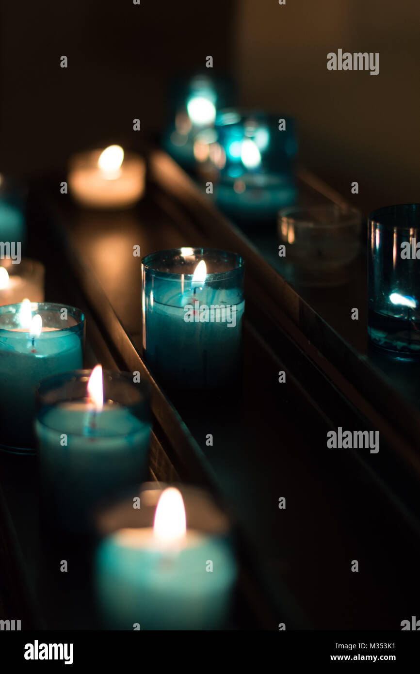 Lit votive candle in turquoise blue holders on a dark metal shelf in low light. Stock Photo