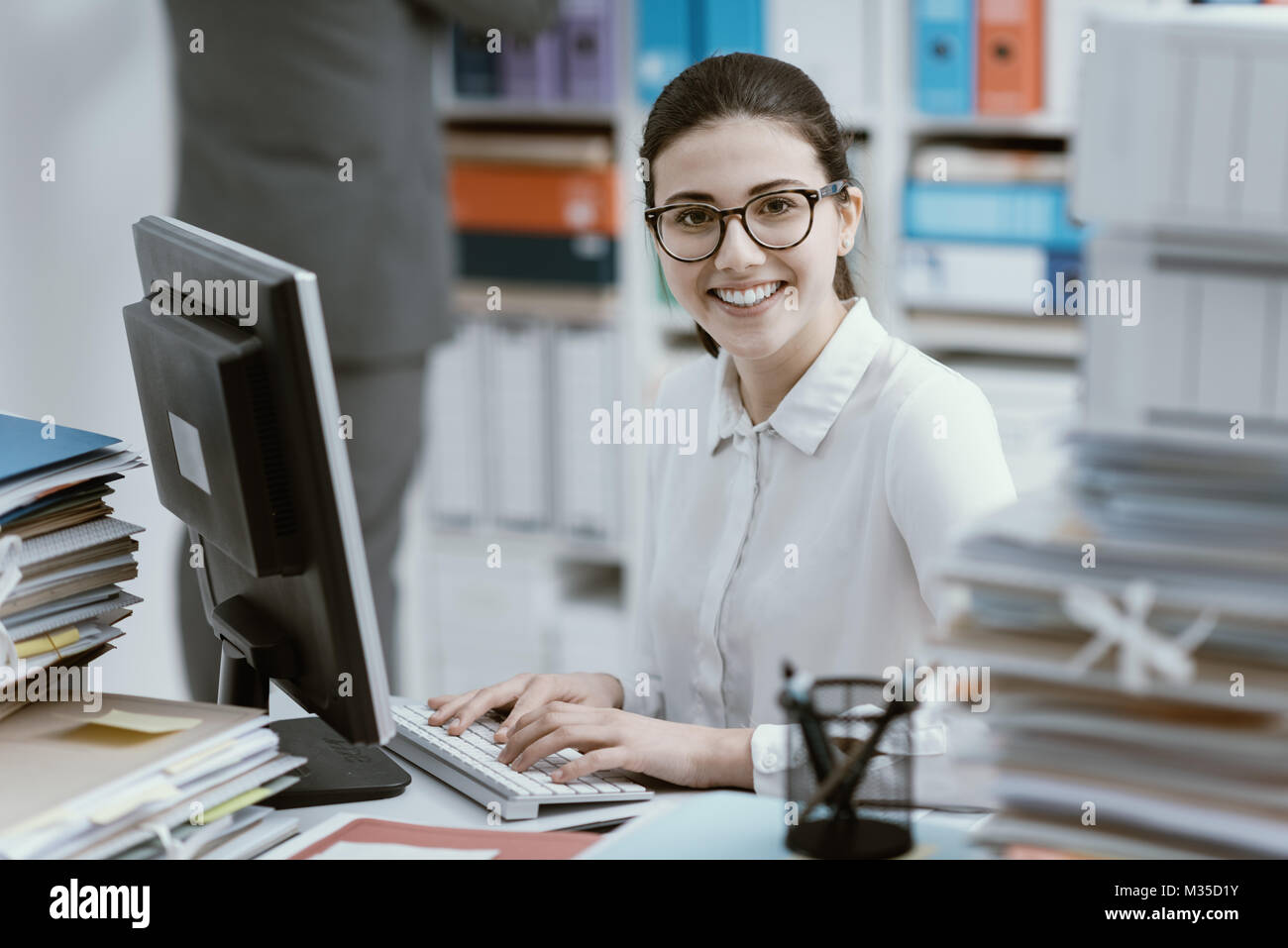 Young smiling secretary working at office desk and stacks of paperwork Stock Photo