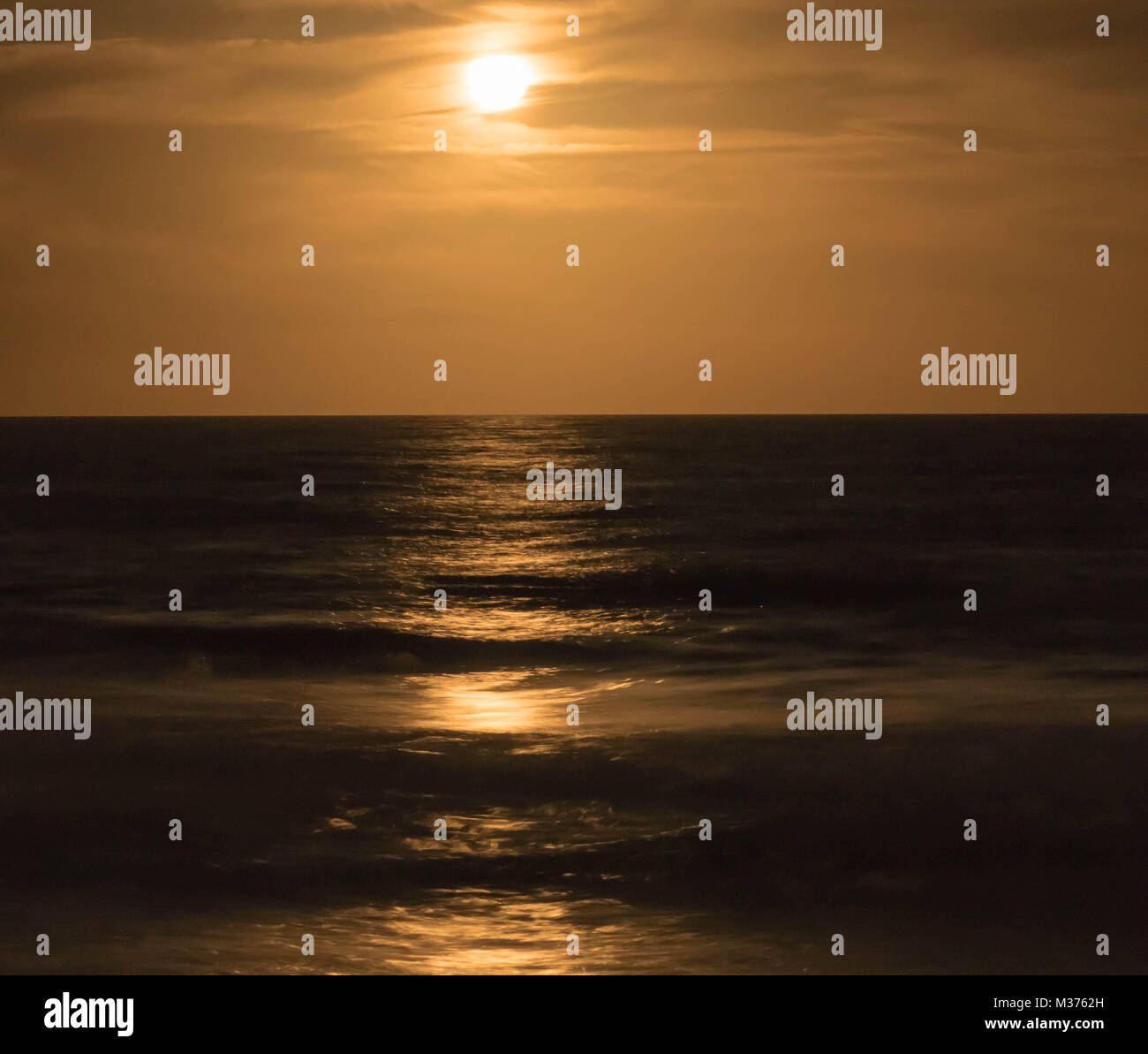 full moon rising into a warm and milky night sky over the ocean Stock Photo
