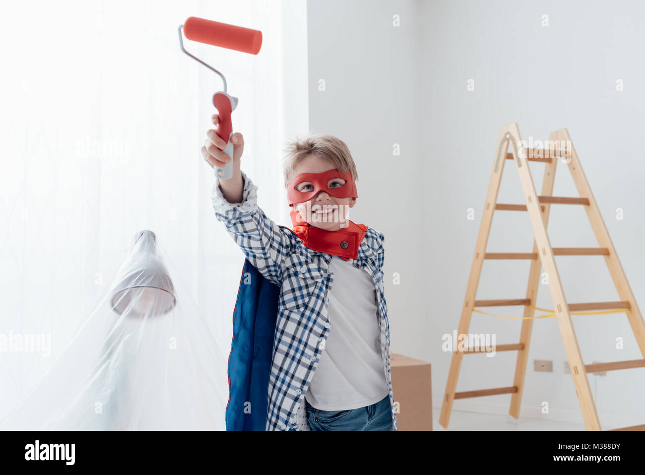 Cute superhero boy with mask and cape, he is holding a paint roller and smiling at camera, home renovation and diy concept Stock Photo