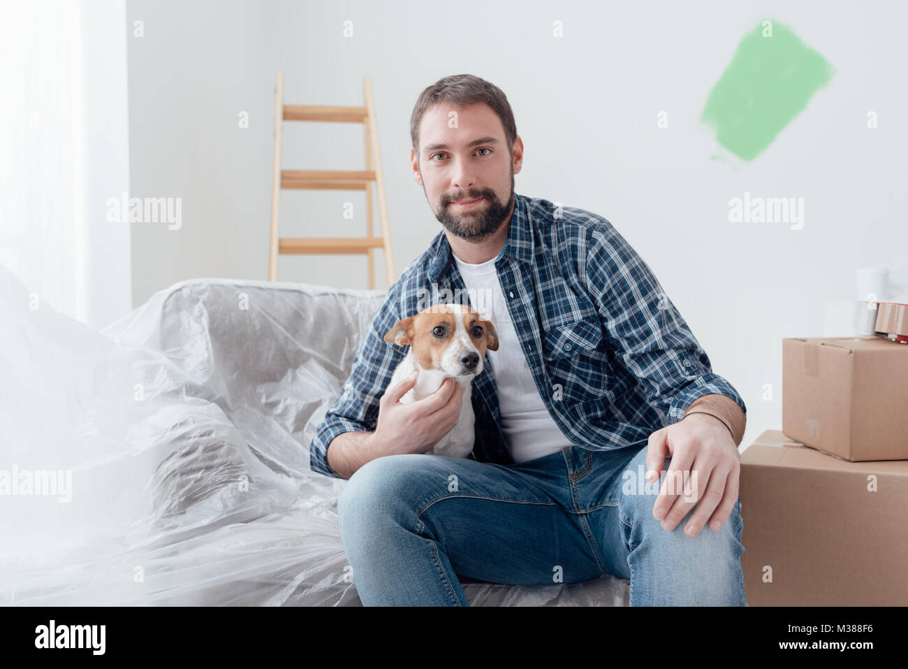 Happy dog owner posing with his pet in his new house during renovation, boxes and ladder on the background Stock Photo