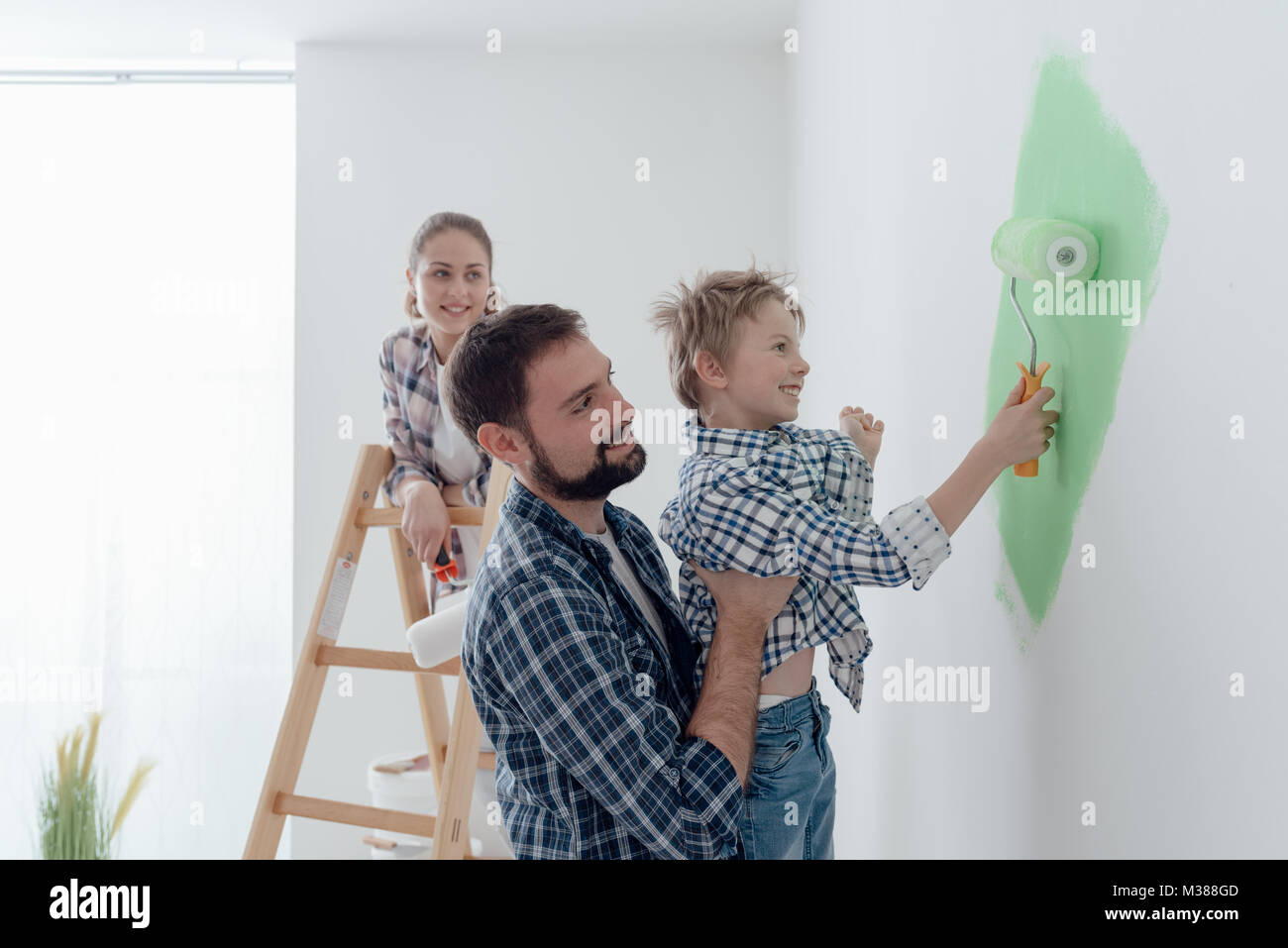 Happy young family renovating their home, the father is holding his son and he is helping him to paint a wall with a paint roller, the mother is stand Stock Photo