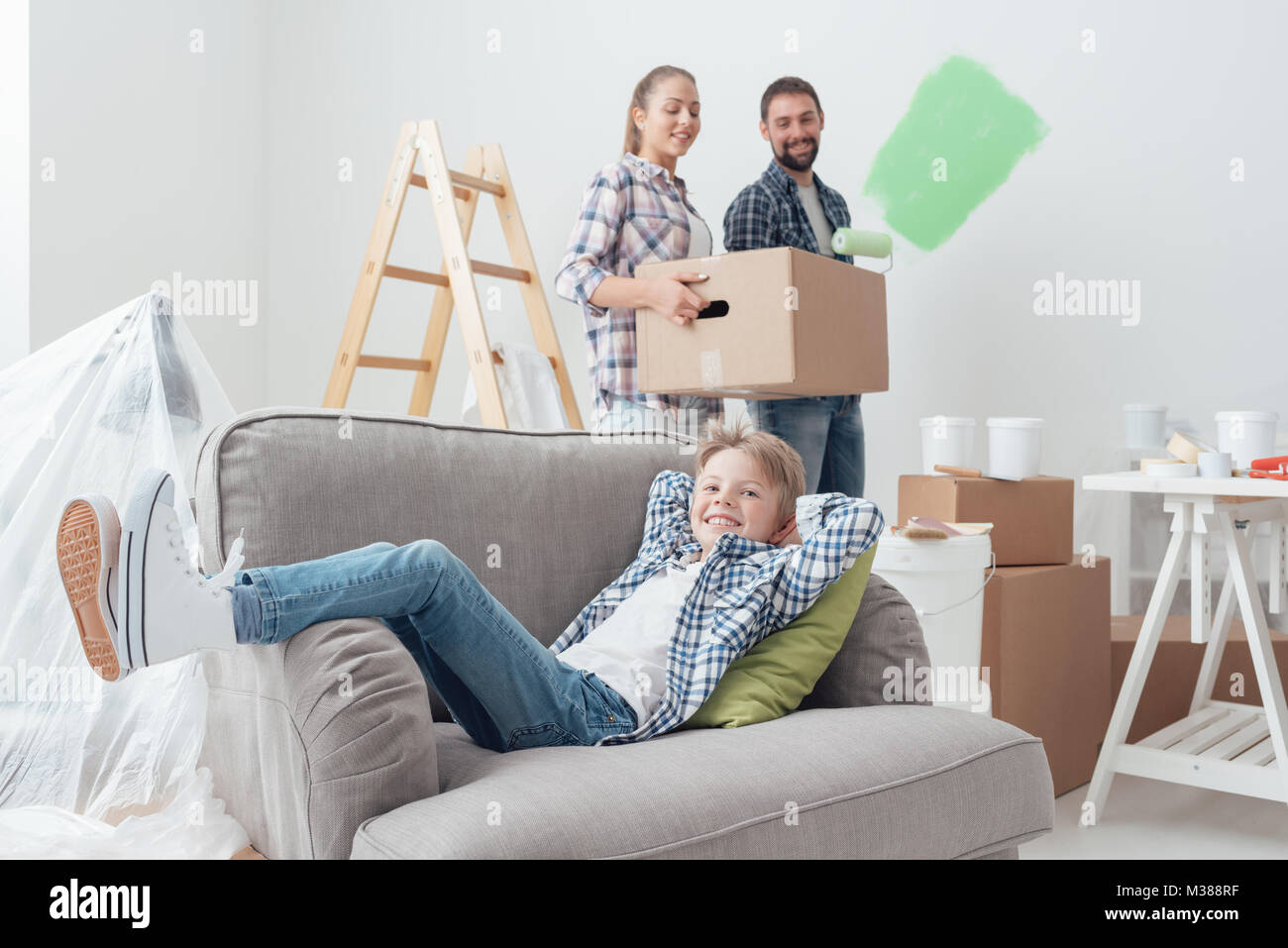 Young family moving into a new house, the mother is carrying a box, the father is painting the room and the child is relaxing on the armchair Stock Photo