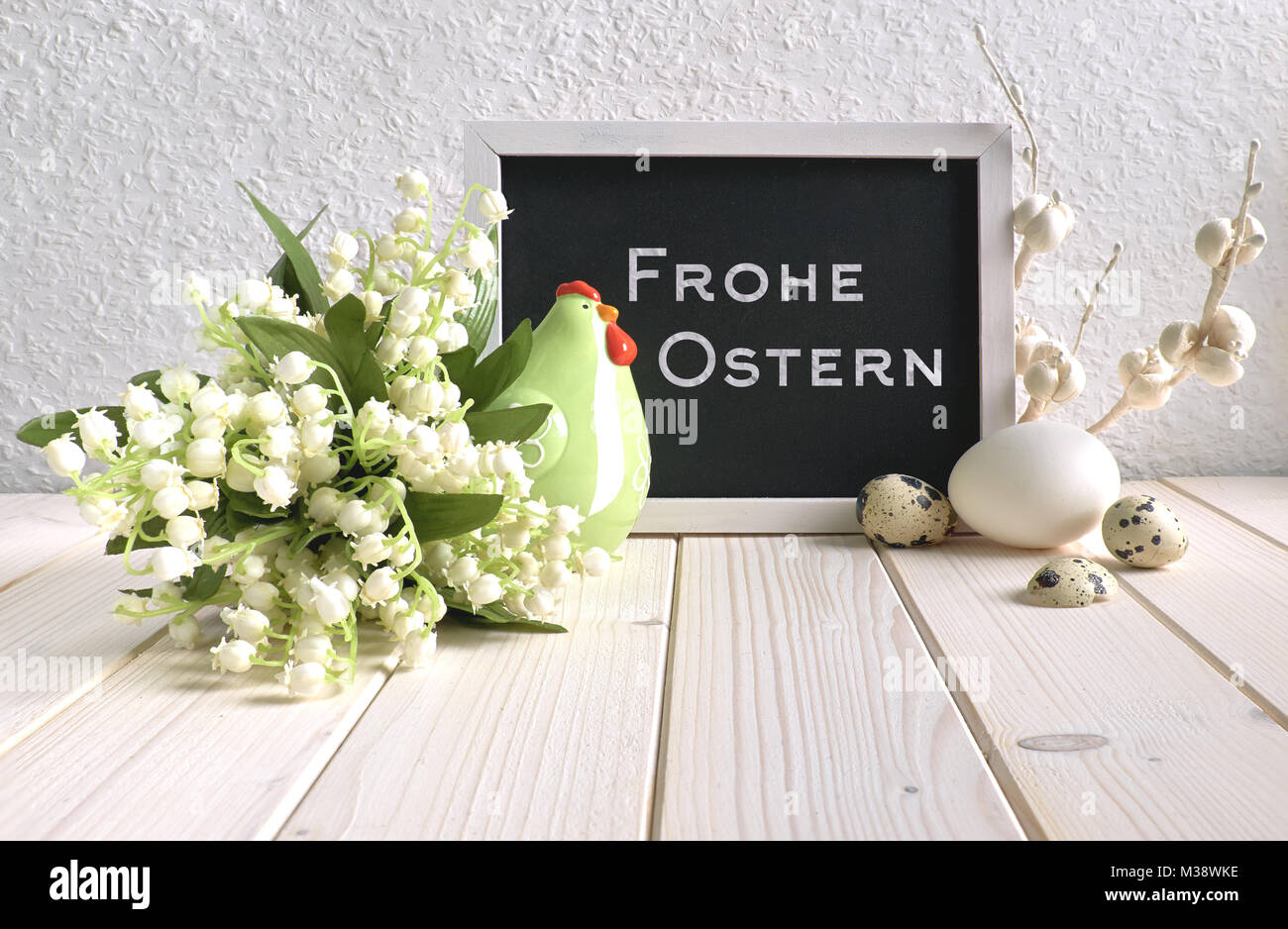 Easter composition with blackboard decorated with ceramic hen, eggs and and lily of the valley flowers, caption 'Frohe Ostern' means 'Happy Easter' in Stock Photo