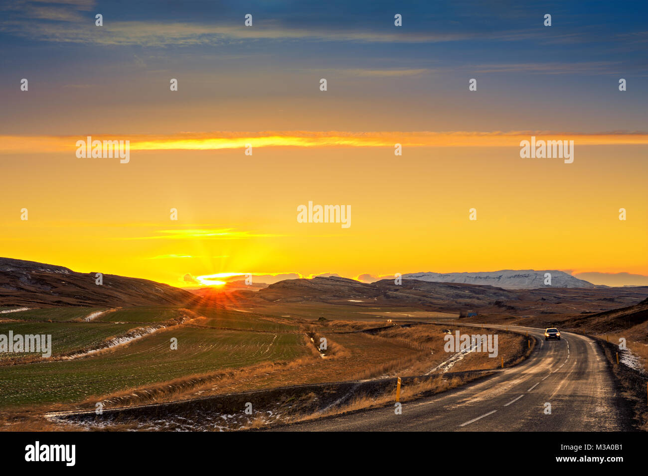Winding road with lonely car crosses farm fields and mountains in rural Iceland at sunset. Stock Photo