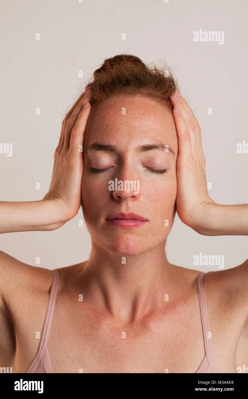 MODEL RELEASED. Close-up of woman covering ears with eyes closed against grey backdrop in studio. Stock Photo