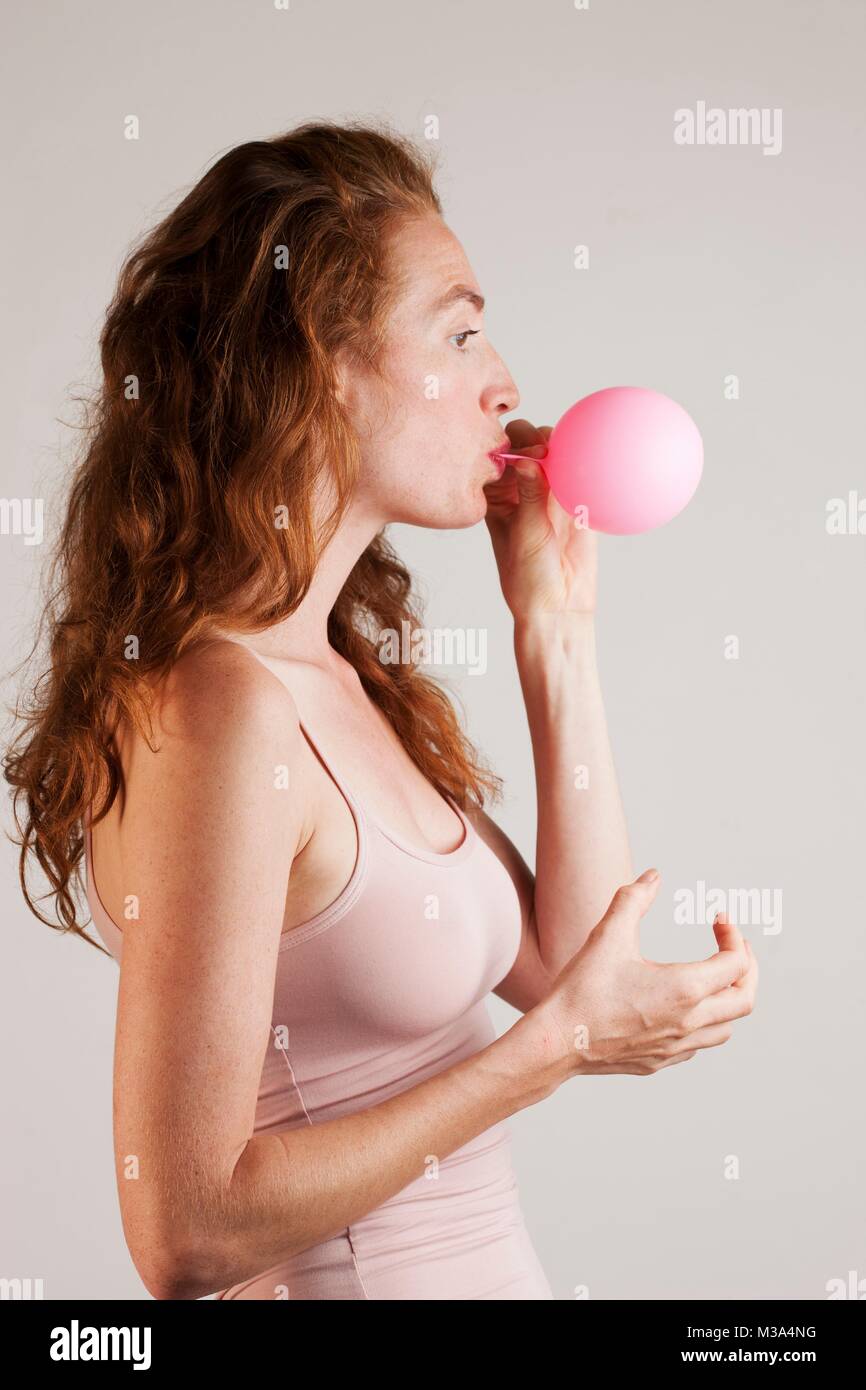 MODEL RELEASED. Profile of young woman with red hair blowing up pink balloon. Stock Photo