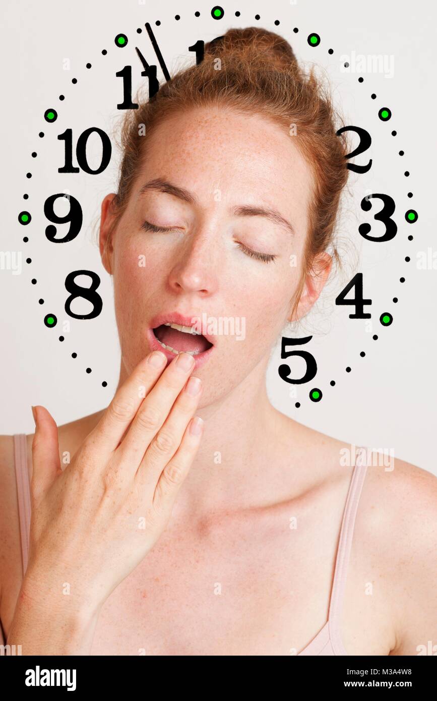 MODEL RELEASED. Young, tired woman yawning and covering her mouth with clock in the background. Stock Photo
