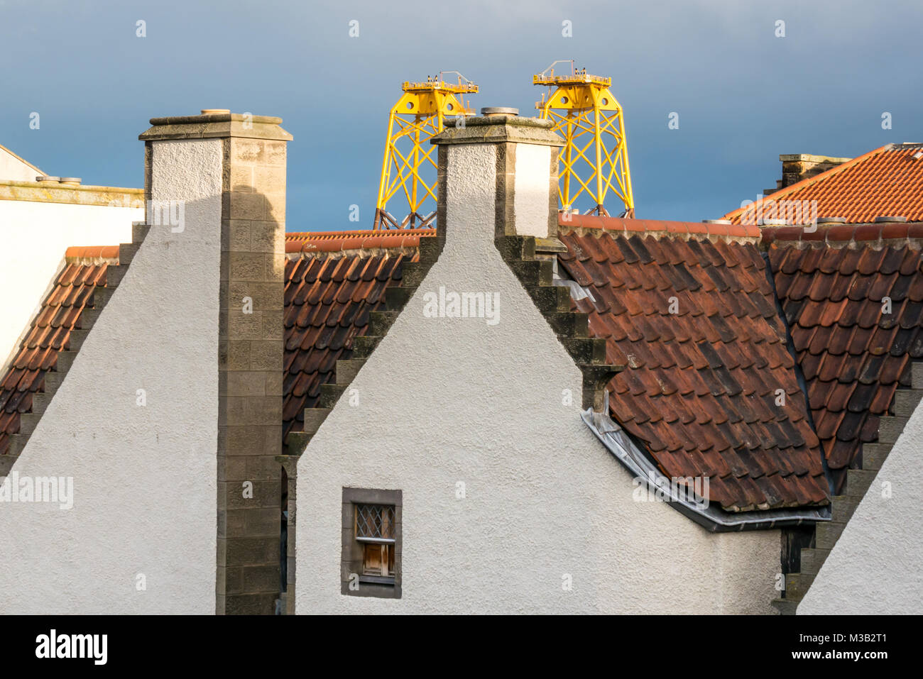 Leith, Edinburgh, Scotland, United Kingdom. Incongruous juxtaposition of huge yellow sunlit wind turbine platforms, called windfarm jackets, in Leith harbour towering over the pantile roof of one of the oldest buildings in Leith, 17th century Lamb's House, a former Hanseatic Leith Merchant house, now home to the architect who restored it, Nicholas Groves Raines. These platforms arrive in Leith on their way North to provide a sub structure for turbines in an offshore wind farm in the Moray Firth Stock Photo