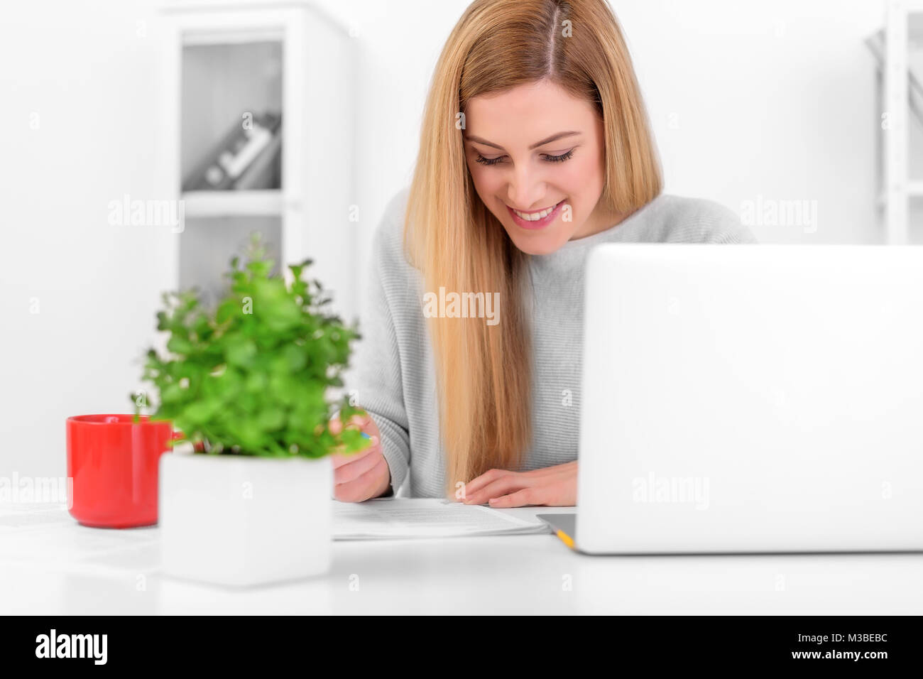 Attractive young woman in a good mood works with documents in the office or at home. Writes, fills papers at the desk with a laptop. Stock Photo