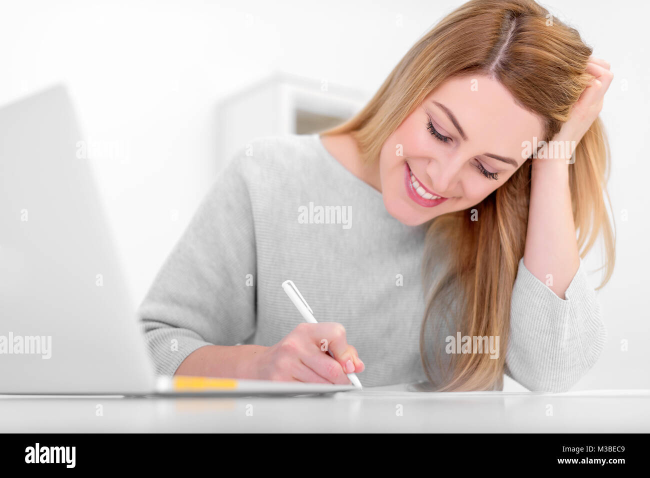 Attractive young woman in a good mood works with documents in the office or at home. Writes, fills papers at the desk with a laptop. Stock Photo