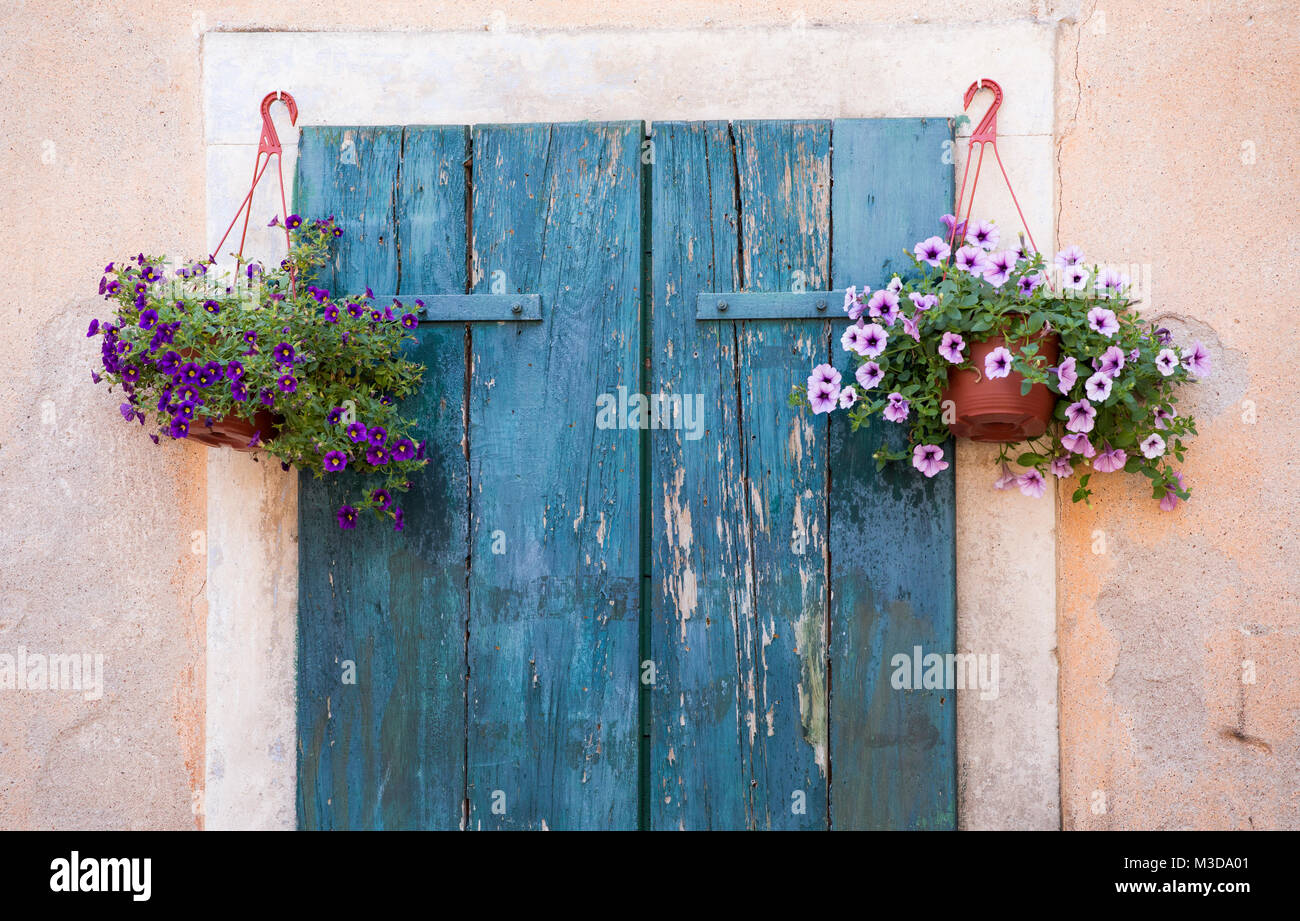 A photograph of weathered wooden shutters in Montenegro, bordered by tow hanging baskets filled with colourful flowers Stock Photo