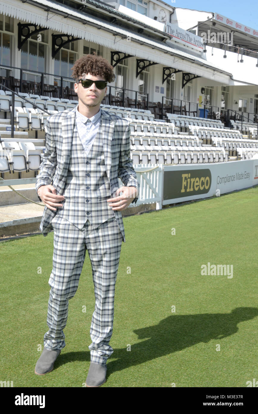 A handsome young mixed race man wearing a patterned suit and sunglasses with afro hair stands at the edge of a Cricket pitch with stalls behind him. Stock Photo
