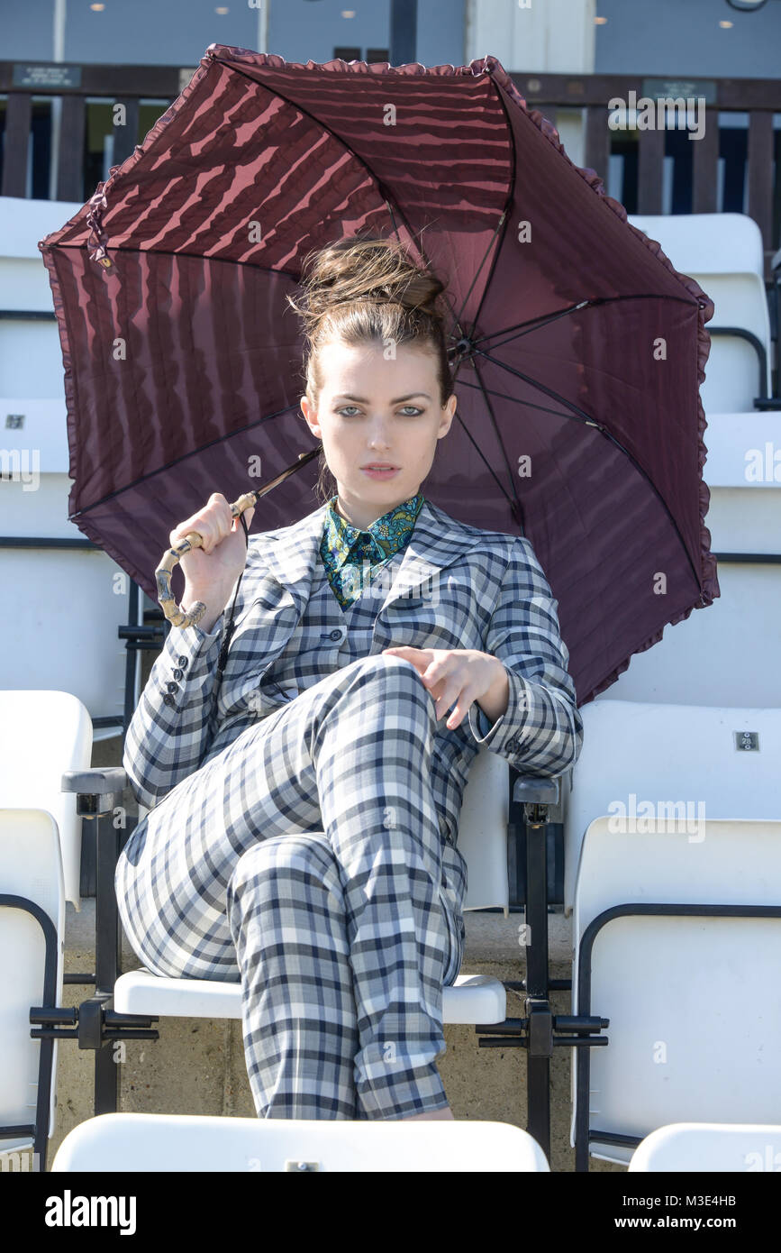 A beautiful girl wearing a tailored stylish patterned suit is at a Cricket Ground on a beautiful bright day- she has a red parasole Stock Photo