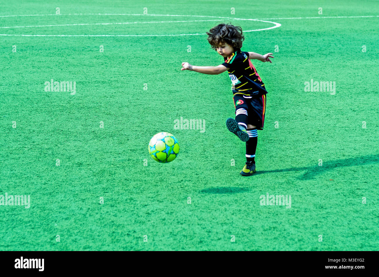 A cute, curly-haired boy kicks a soccer ball on a field of green turf. Stock Photo