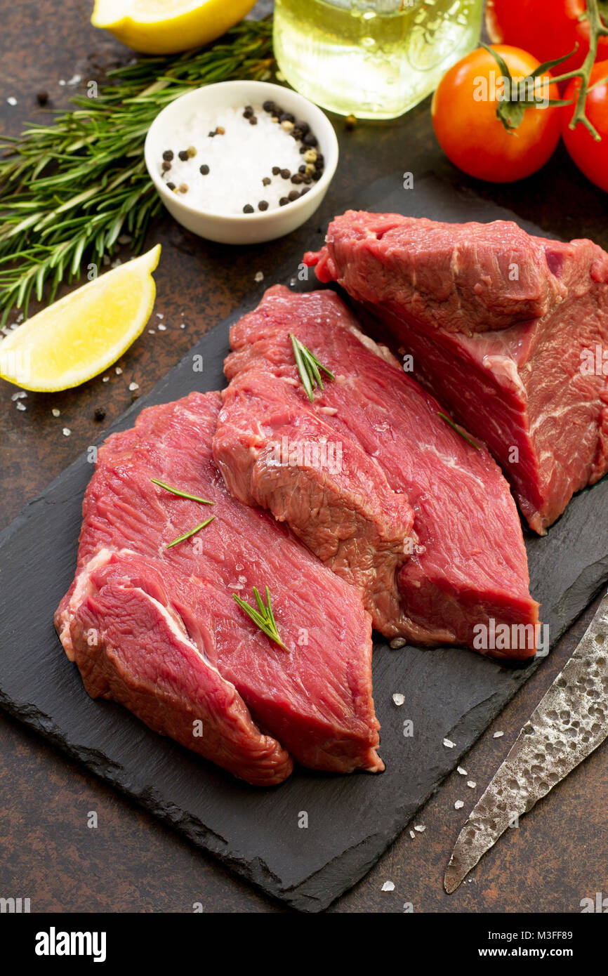 Fresh meat. Cheese steak beef on a slate cutting board, lemon, spices and fresh rosemary on a stone table. Stock Photo