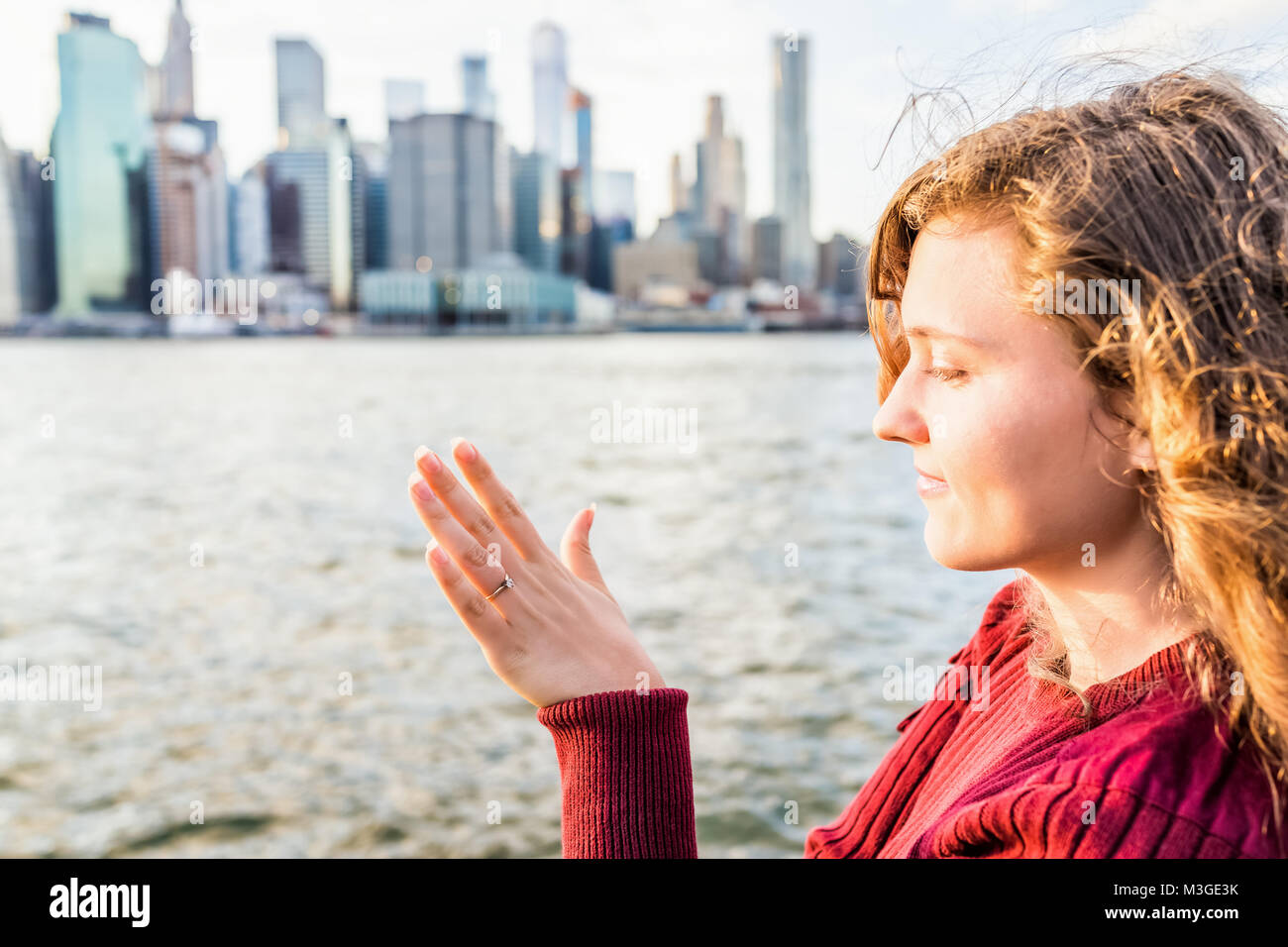 Young woman's hand with diamond engagement ring solitaire, gold wedding band outside outdoors in NYC New York City Brooklyn Bridge Park by east river, Stock Photo
