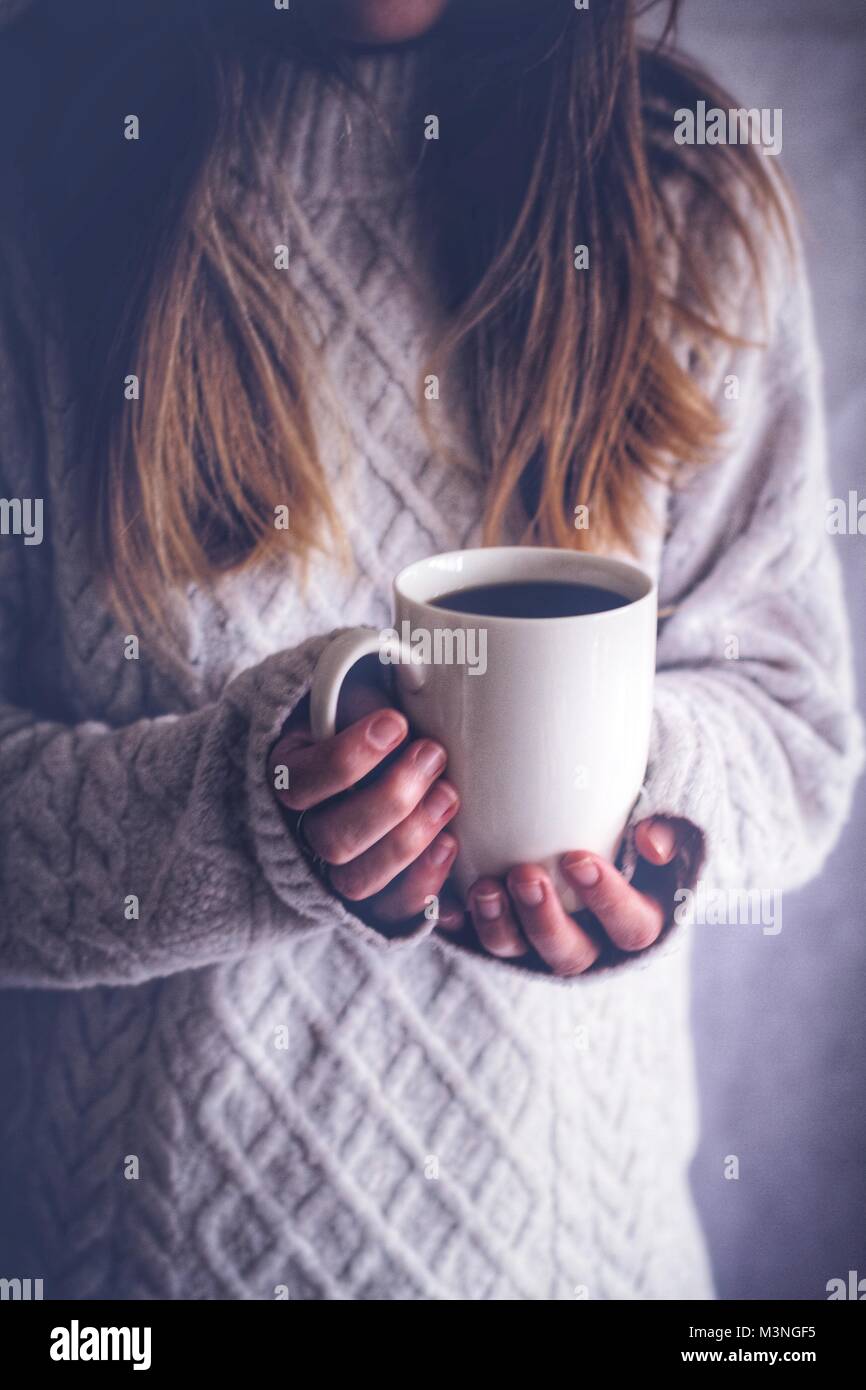 A midsection of a blonde haired girl holding up a white cup indoors and in the shadows. Stock Photo