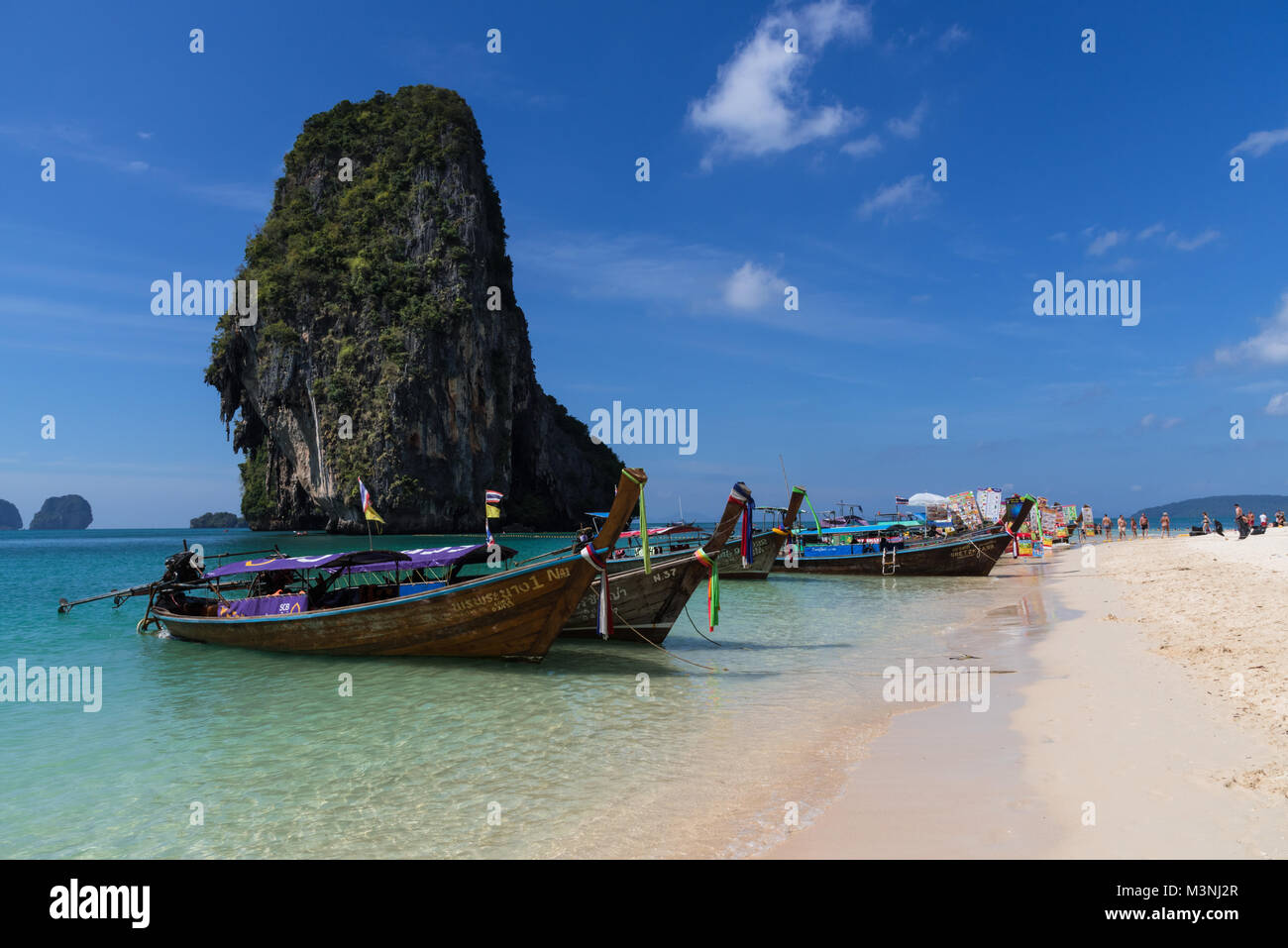 Stunning landscape view of a row of long-tail boats anchored on a beautiful golden sandy beach in Thailand with blue sky and towering rock island Stock Photo