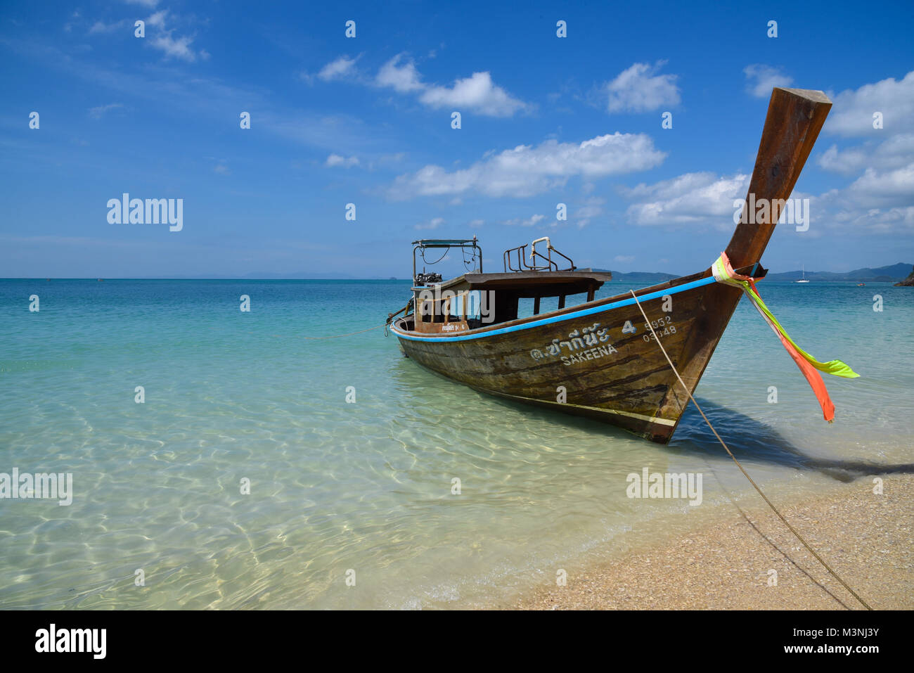 Single long-tail boat anchored on a yellow sandy beach with beautiful clear turquoise blue sea, blue sky, light white cloud in Thailand Stock Photo