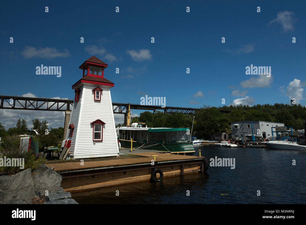 Waterfront lighthouse on dock, train trestle, boats sunny blue sky in background Summer in Georgian Bay National Park Parry Sound Ontario Canada Stock Photo