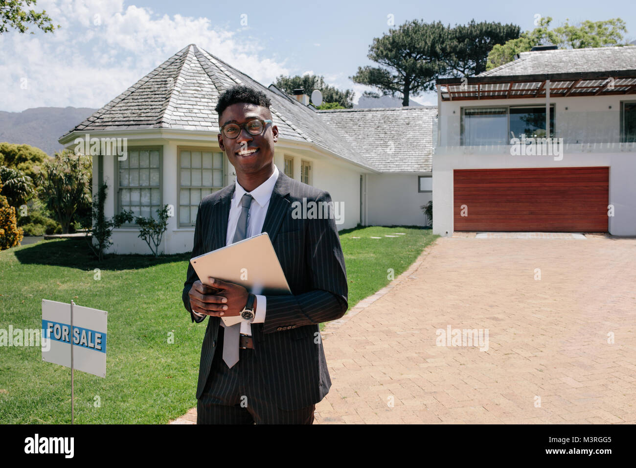 Portrait of confident young african male real estate agent standing outside a house for sale. Realtor with digital tablet waiting for visitors. Stock Photo