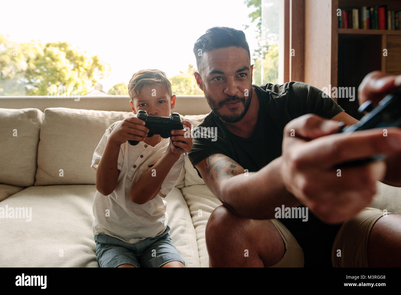 Father and son playing video game at home. Young man and little boy sitting on sofa in living room and playing video game. Stock Photo