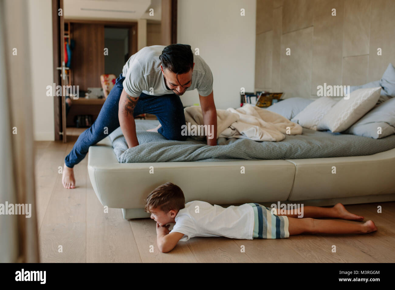 Father and son playing hide and seek in bedroom. Little boy hiding by the bed with father searching him. Family playing games inside their home. Stock Photo