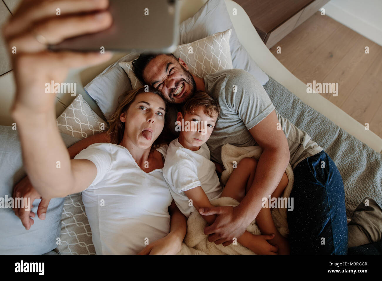 Happy young woman sticking out her tongue and taking selfie with her crazy family. Family lying on bed with funny facial expression and taking selfie  Stock Photo