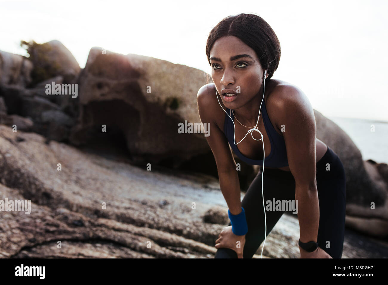 Tired young woman resting after running outdoors. African female runner standing with hands on knees. Stock Photo