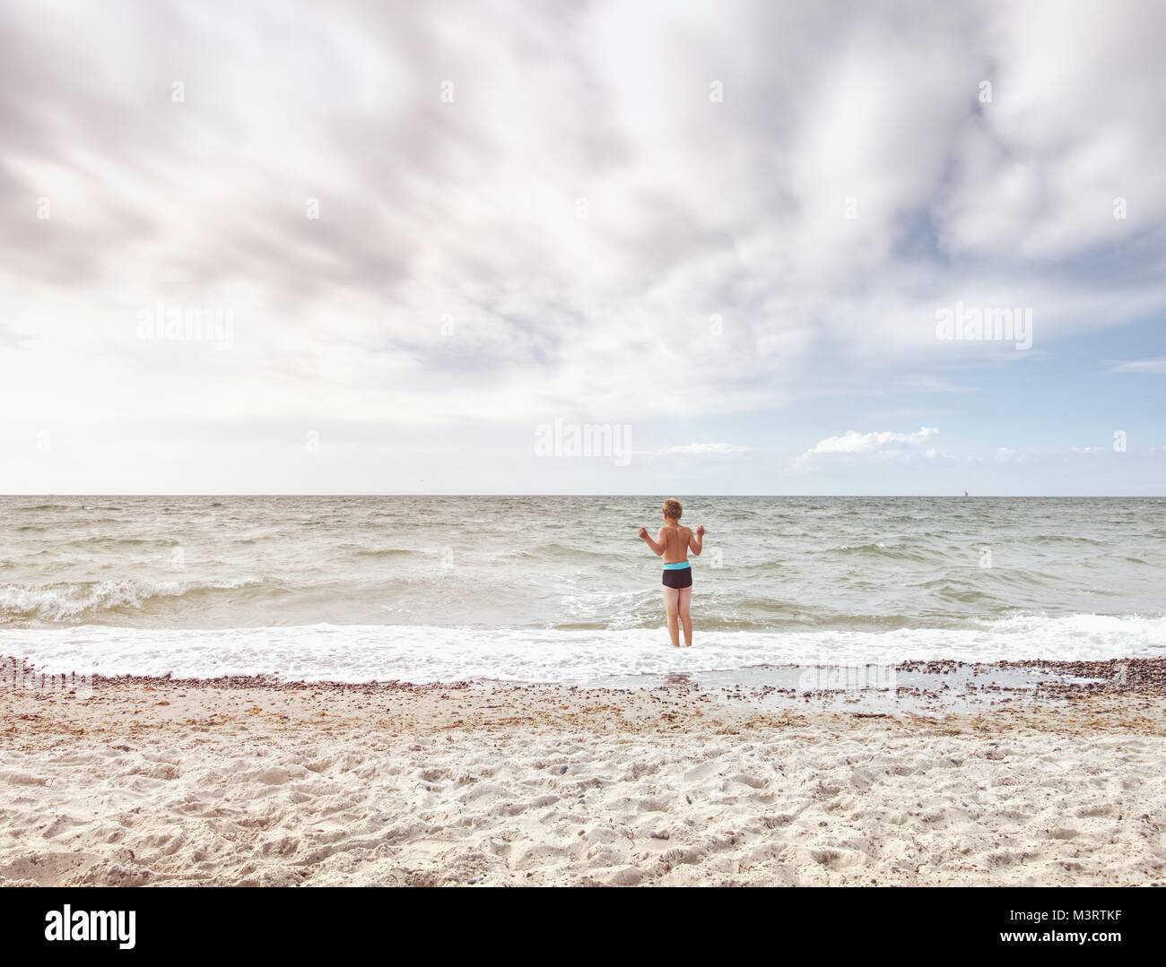 Blond hair boy stay in cold sea tide. Kid on stony beach with foamy waves.  Windy day, cloudy  blue sky on seascape Stock Photo