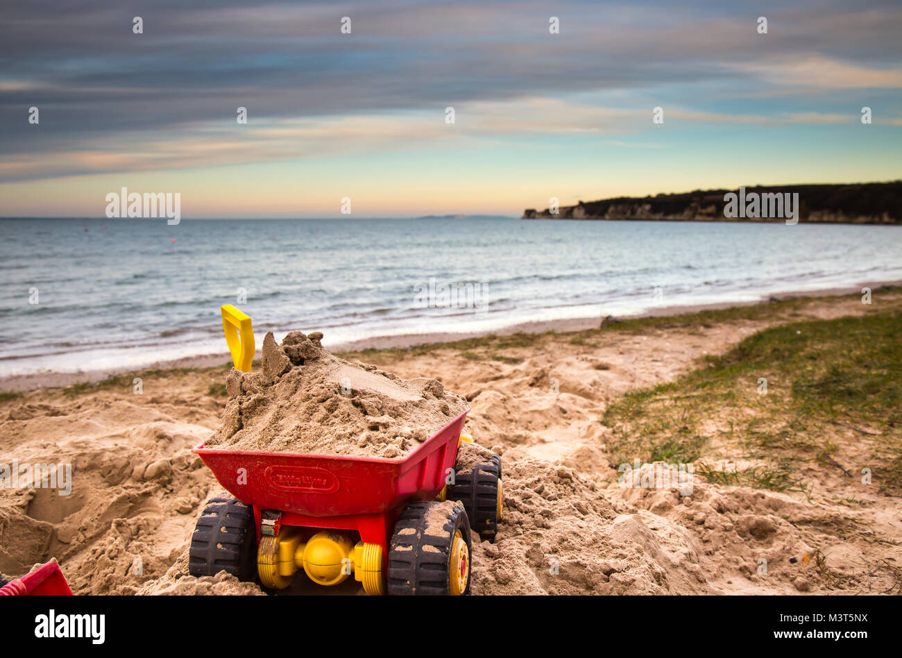 Close up of a child's toy trucks abandoned in the sand, on a deserted UK beach. Early evening sunlight creates stunning colours across sky. Stock Photo