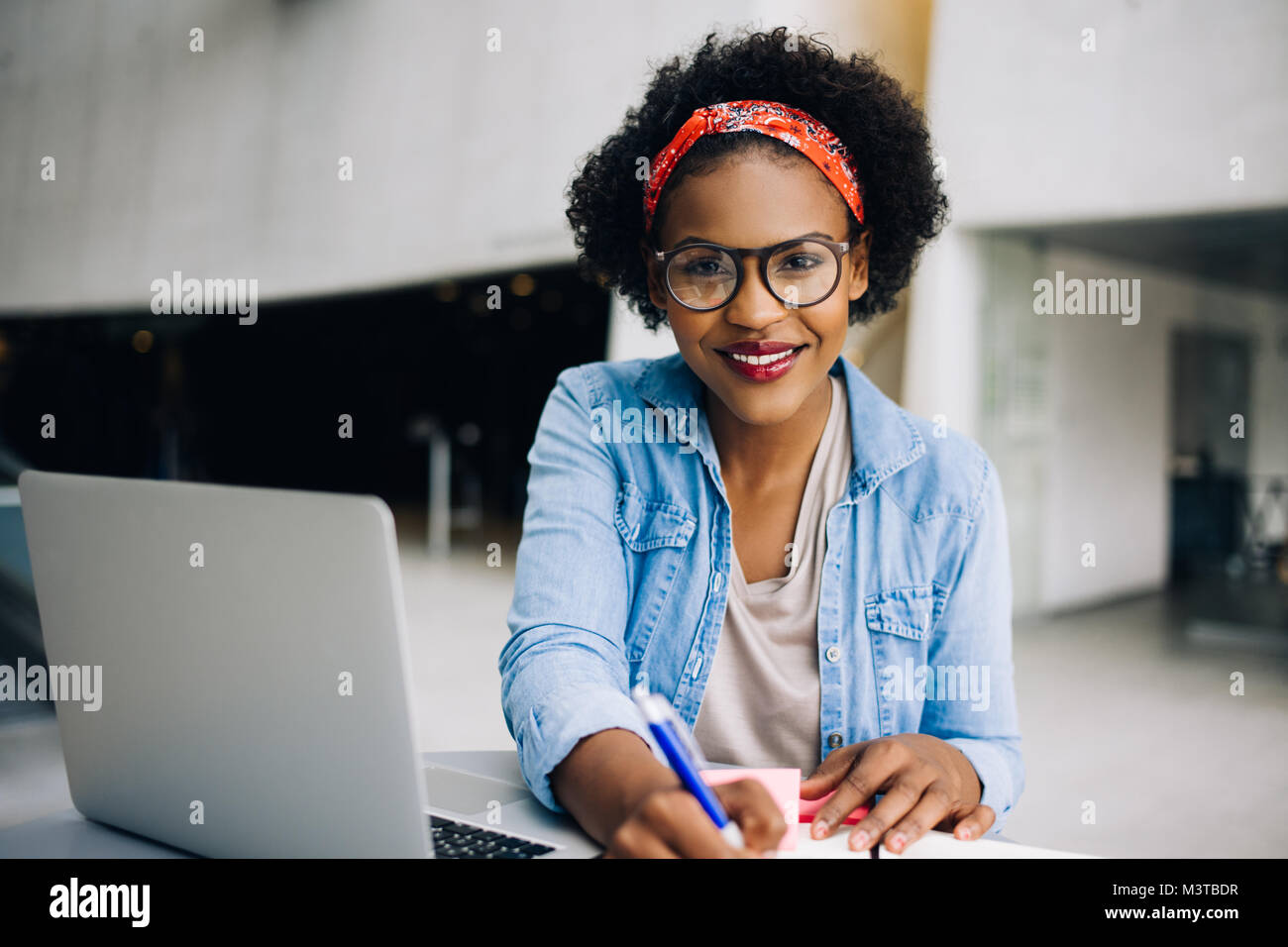 Smiling young African female entrepreneur sitting at a table in a modern office building lobby working on a laptop and writing notes in her planner Stock Photo