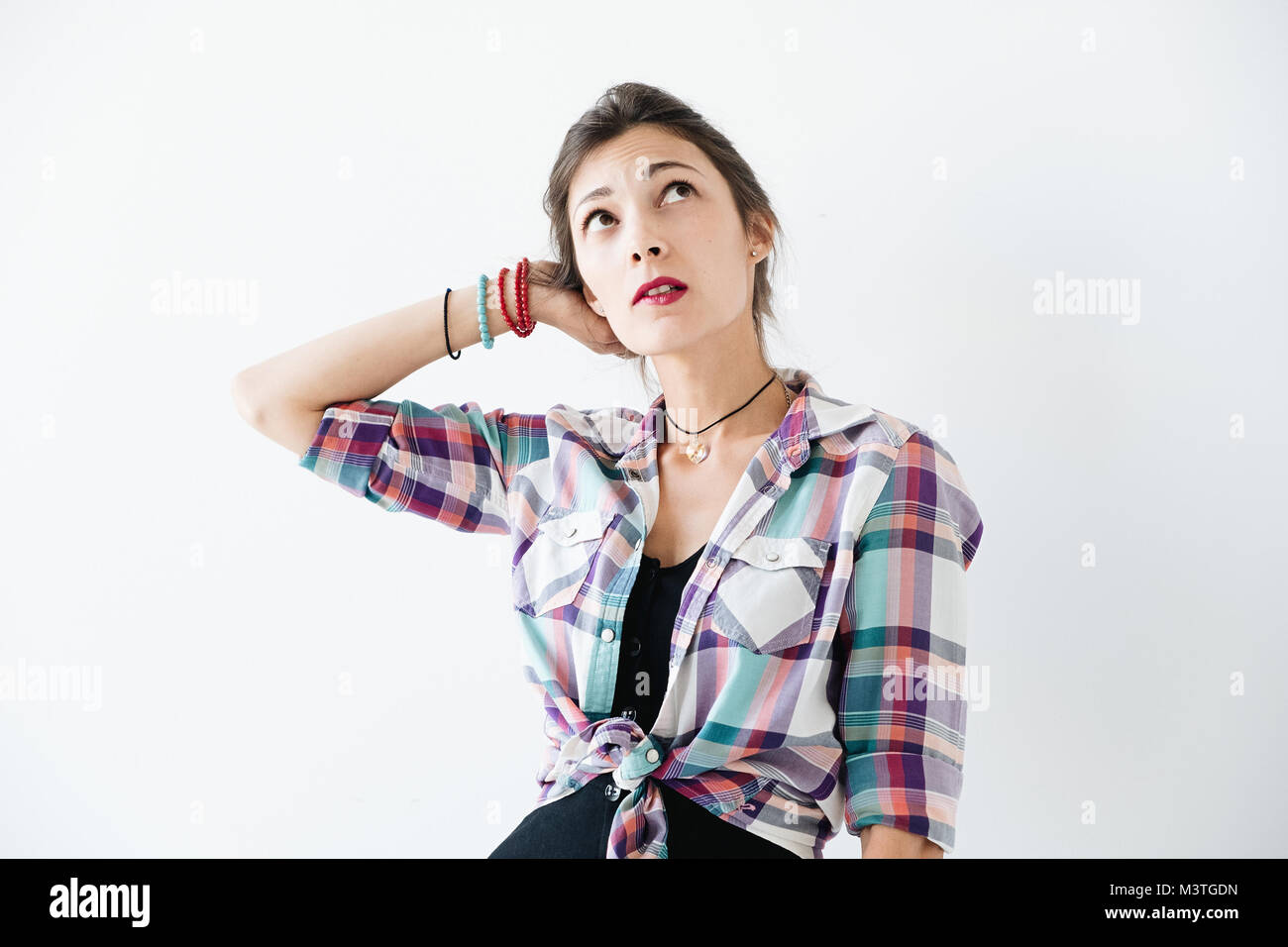 Worried tanned young woman wearing plaid shirt bite her lip and looks up, with copy space above her head. Thoughtful girl holds one arm on her head on Stock Photo