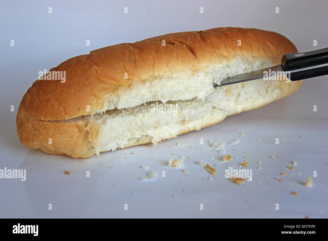 Hot dog bun or small piece of bread being cut with bread knife on clear background with crumbs lying in front Stock Photo