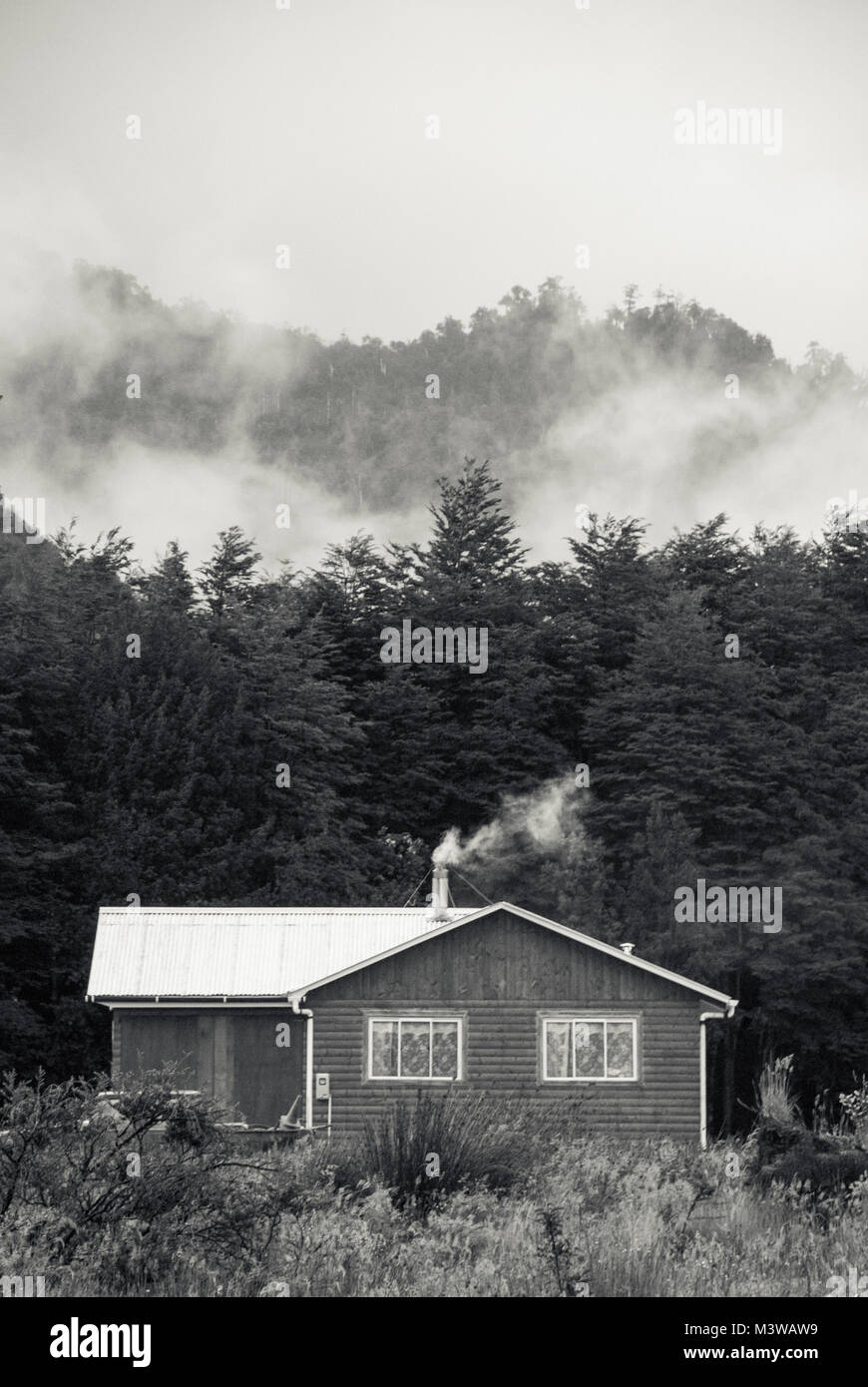 A wooden house with smoke coming from the chimney against a misty forest background Stock Photo