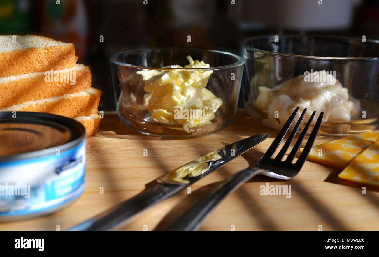 White bread, butter in glass dish, mayo in glass dish, knife and fork, un opened can of tuna fish. Light wood background. Shadowing. Stock Photo