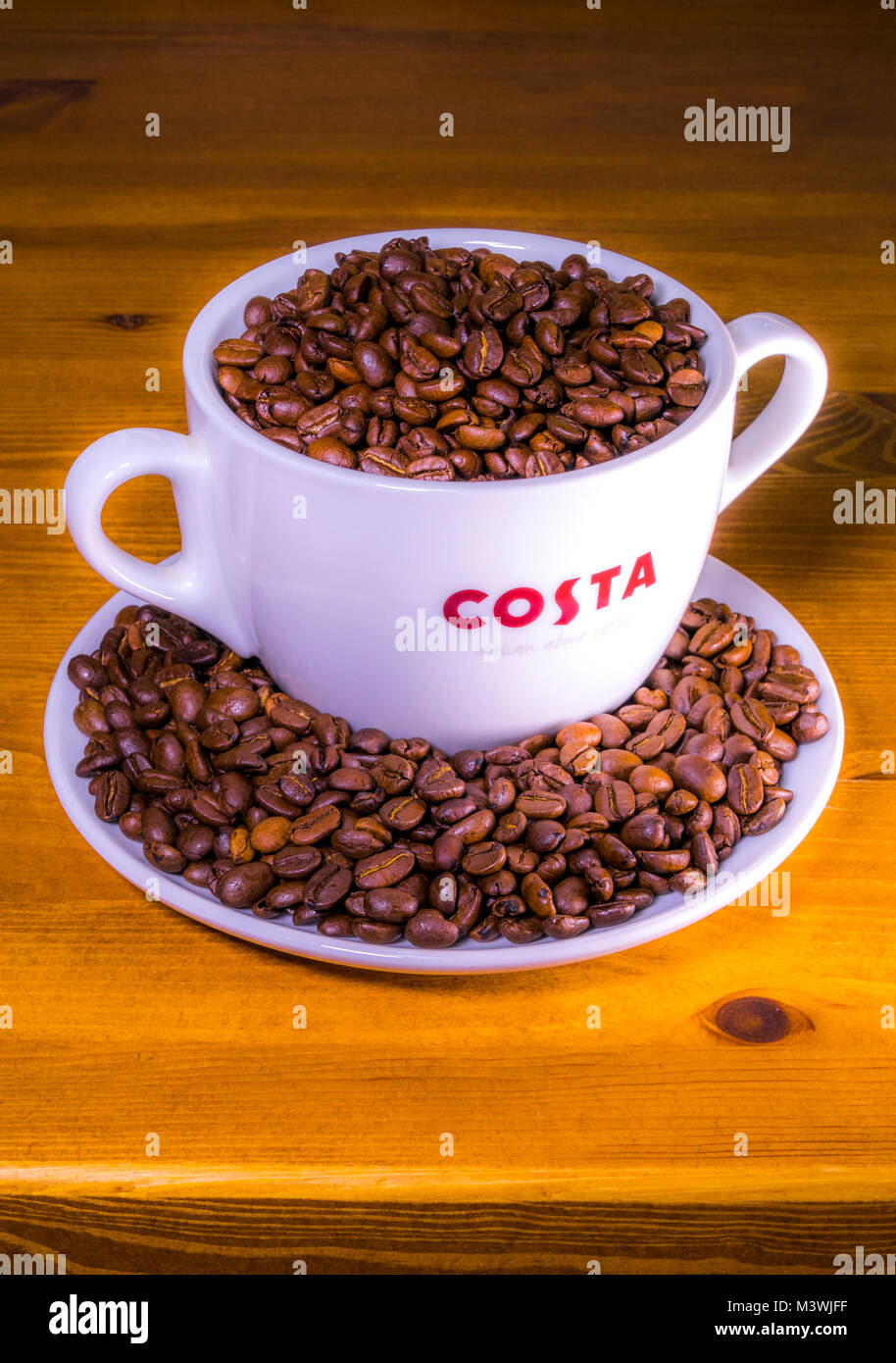 Costa coffee: logo on extra large cup (with dual handles), sitting on saucer, full of roasted whole coffee beans, on an old pine worktop. England, UK. Stock Photo