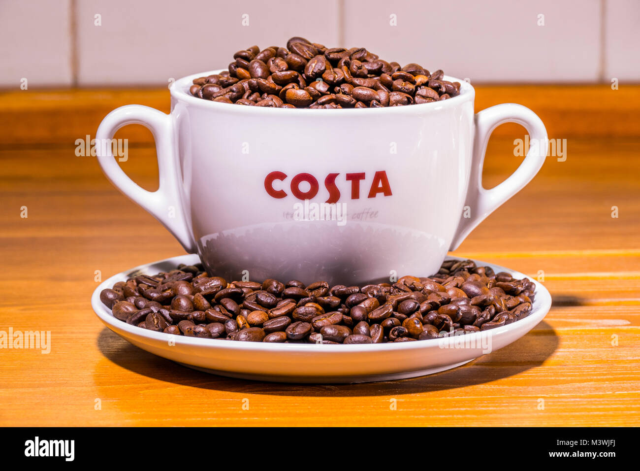 Costa coffee: logo on extra large cup (with dual handles), sitting on saucer, full of roasted whole coffee beans, on an old pine worktop. England, UK. Stock Photo