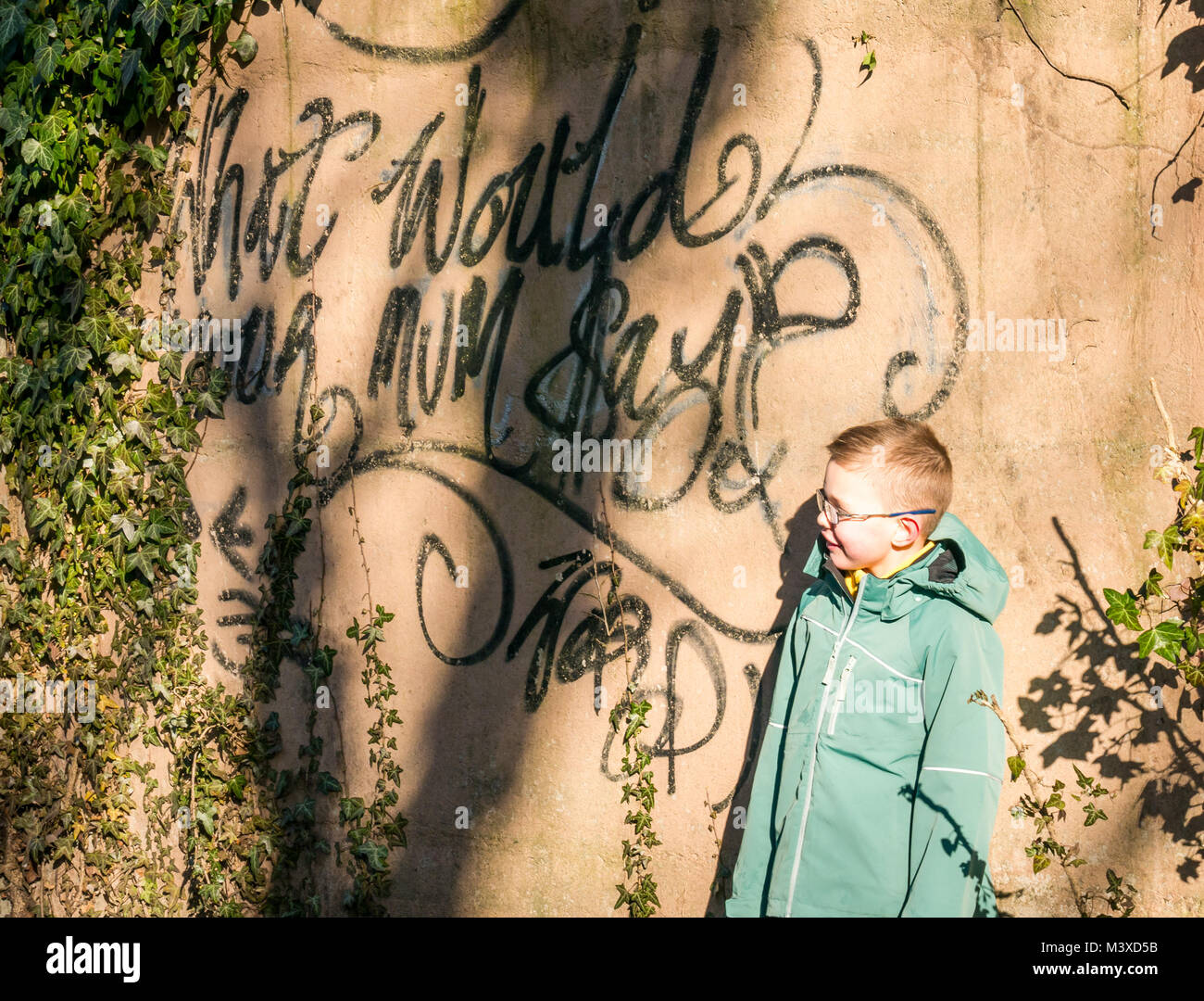 6 year old boy with glasses standing next to spray paint graffiti on wall, which reads What Would Your Mum Say?, Scotland, UK Stock Photo