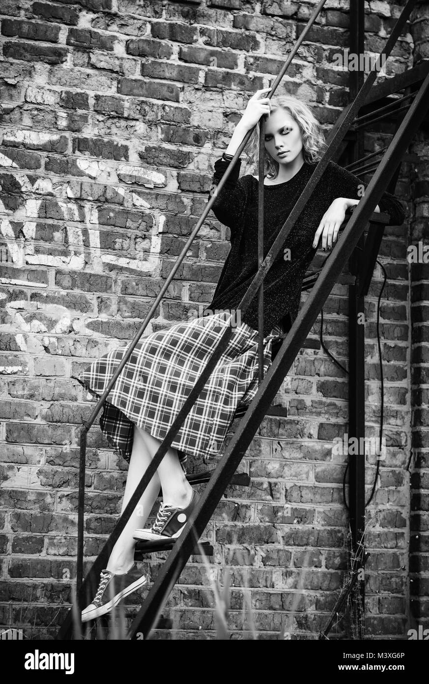 Grunge fashion: cute young girl (informal model) in checkered skirt and jacket sitting on the ladder. Black and white Stock Photo