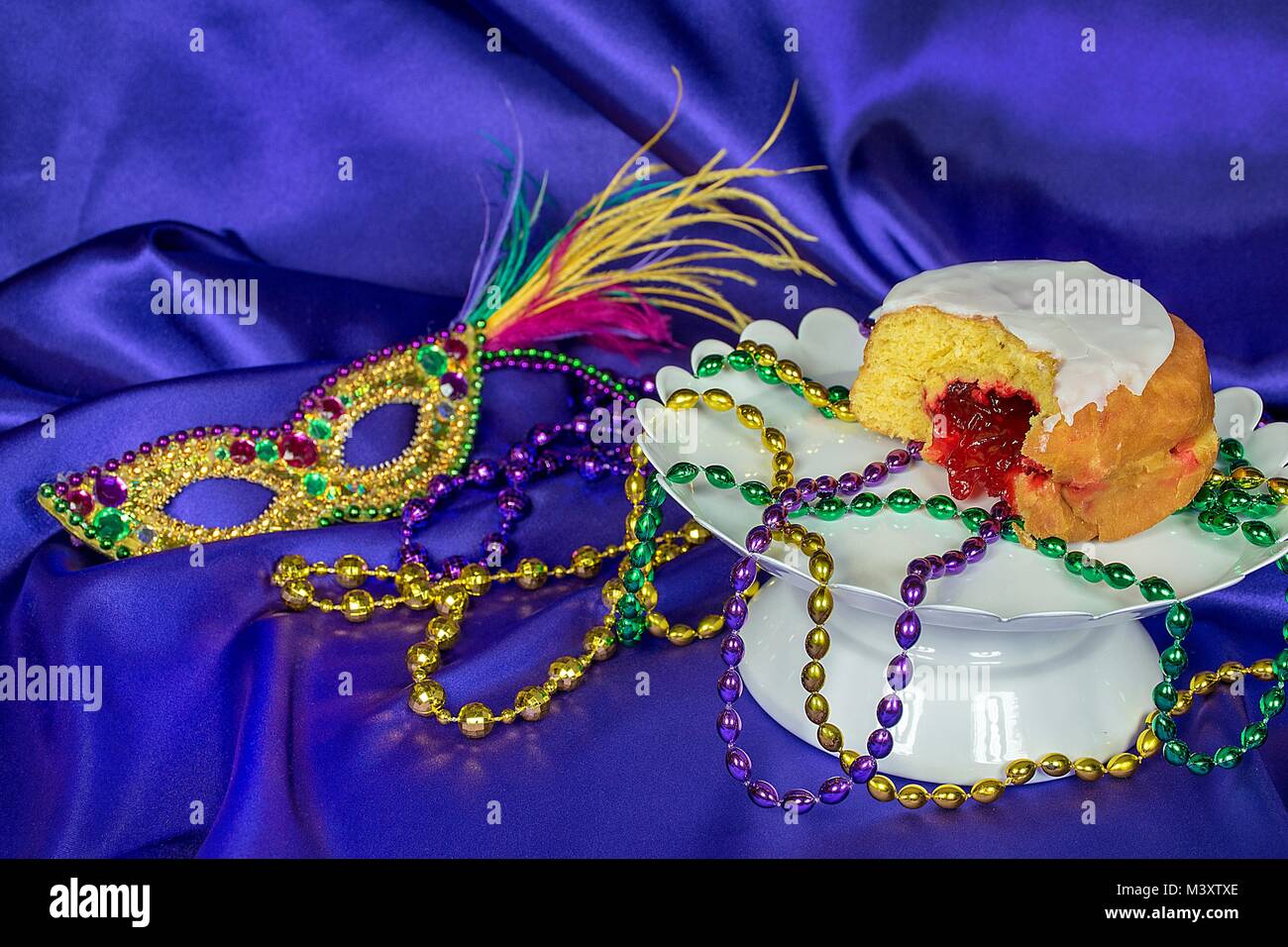 Fat Tuesday paczki with Mardi Gras party beads on white pedestal plate with sequin mask on purple satin Stock Photo