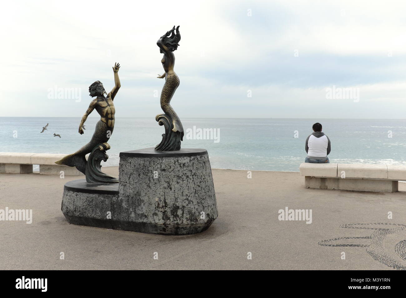 A lone woman sits on the Puerto Vallarta seaside malecon with seagulls flying nearby and a statue of Triton merman and Nereida mermaid behind her. Stock Photo