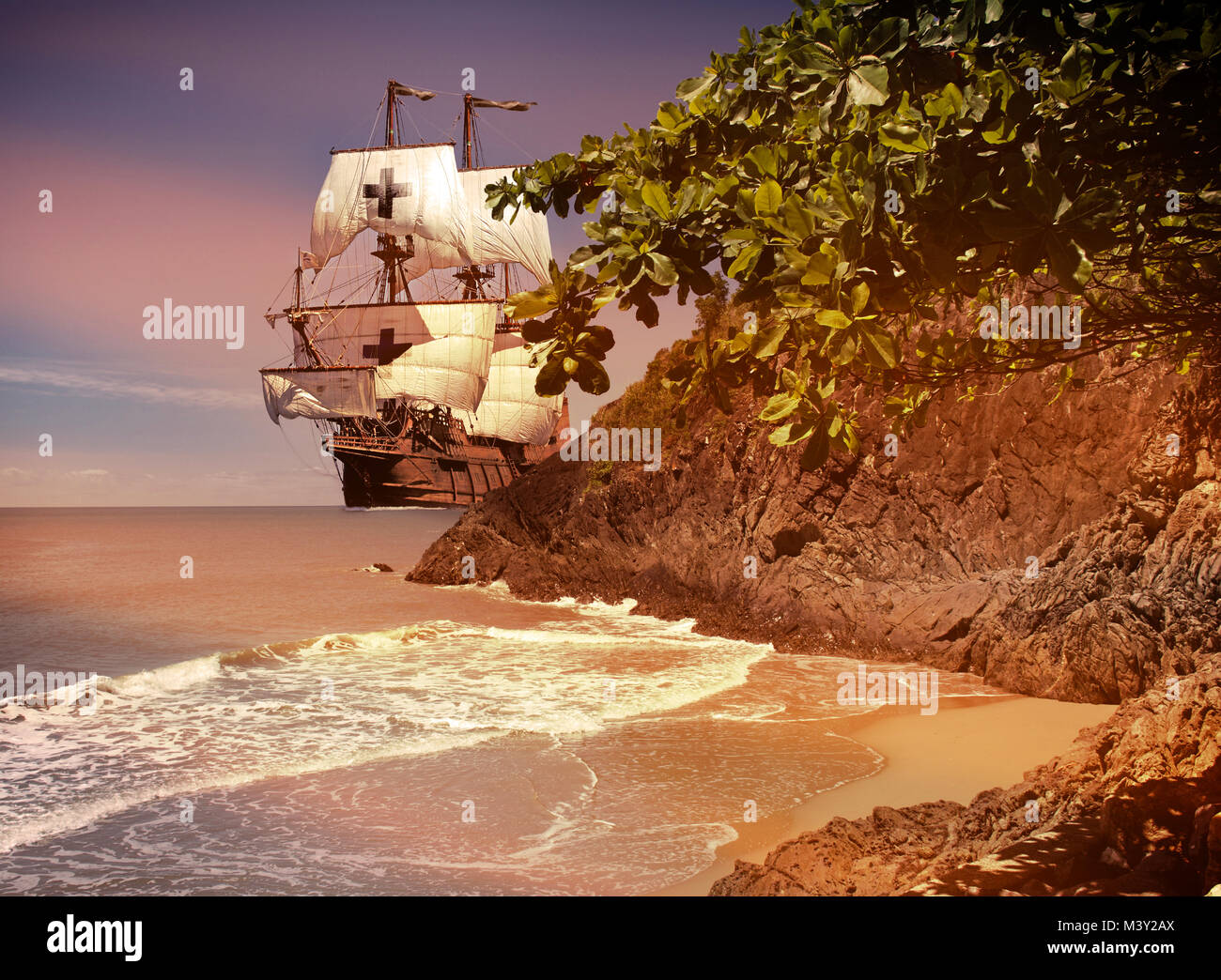 Old wooden tall sail pirate ship rounds a rocky outcrop to a small cove with a sand beach Stock Photo