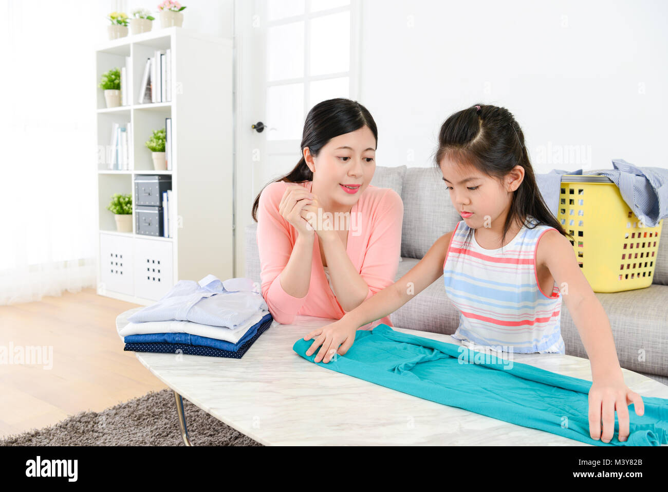 cute attractive little girl learning folding clothing when mother looking at her and explaining organized method for her. Stock Photo