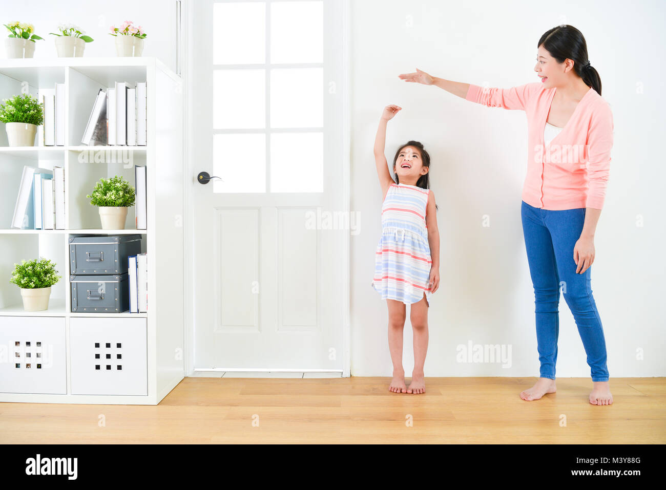 pretty attractive mother with cute little daughter measuring growth record when they standing in wooden floor with white background together. Stock Photo