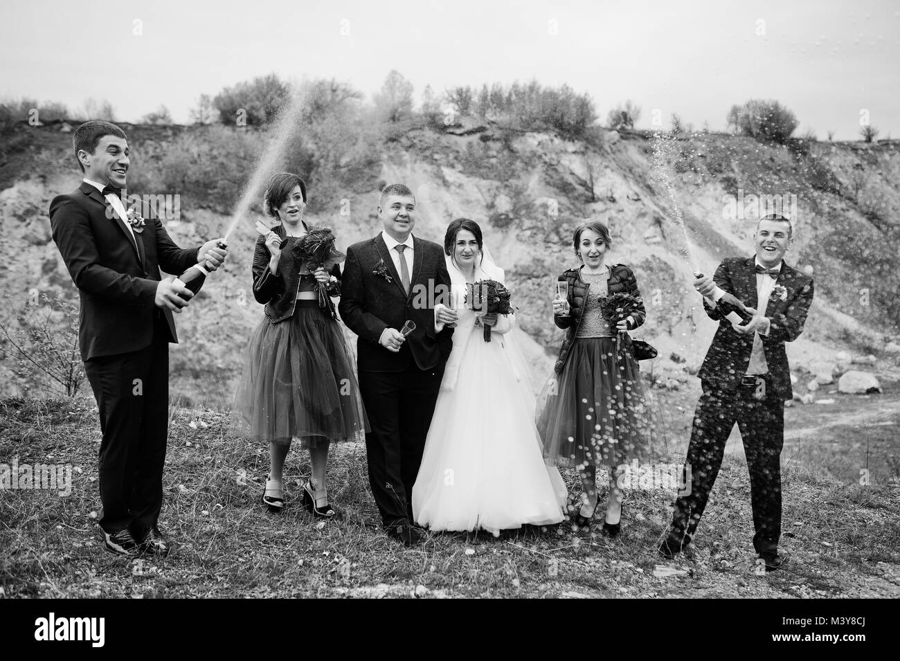 Wedding couple, groomsman and bridesmaids looking at best man opening the champagne bottle in sand quarry. Black and white photo. Stock Photo